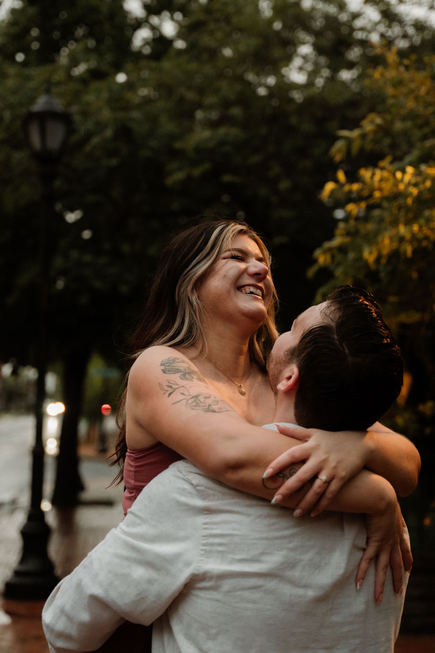 A joyful woman with tattoos laughs while being lifted by her partner outdoors near a street lamp.