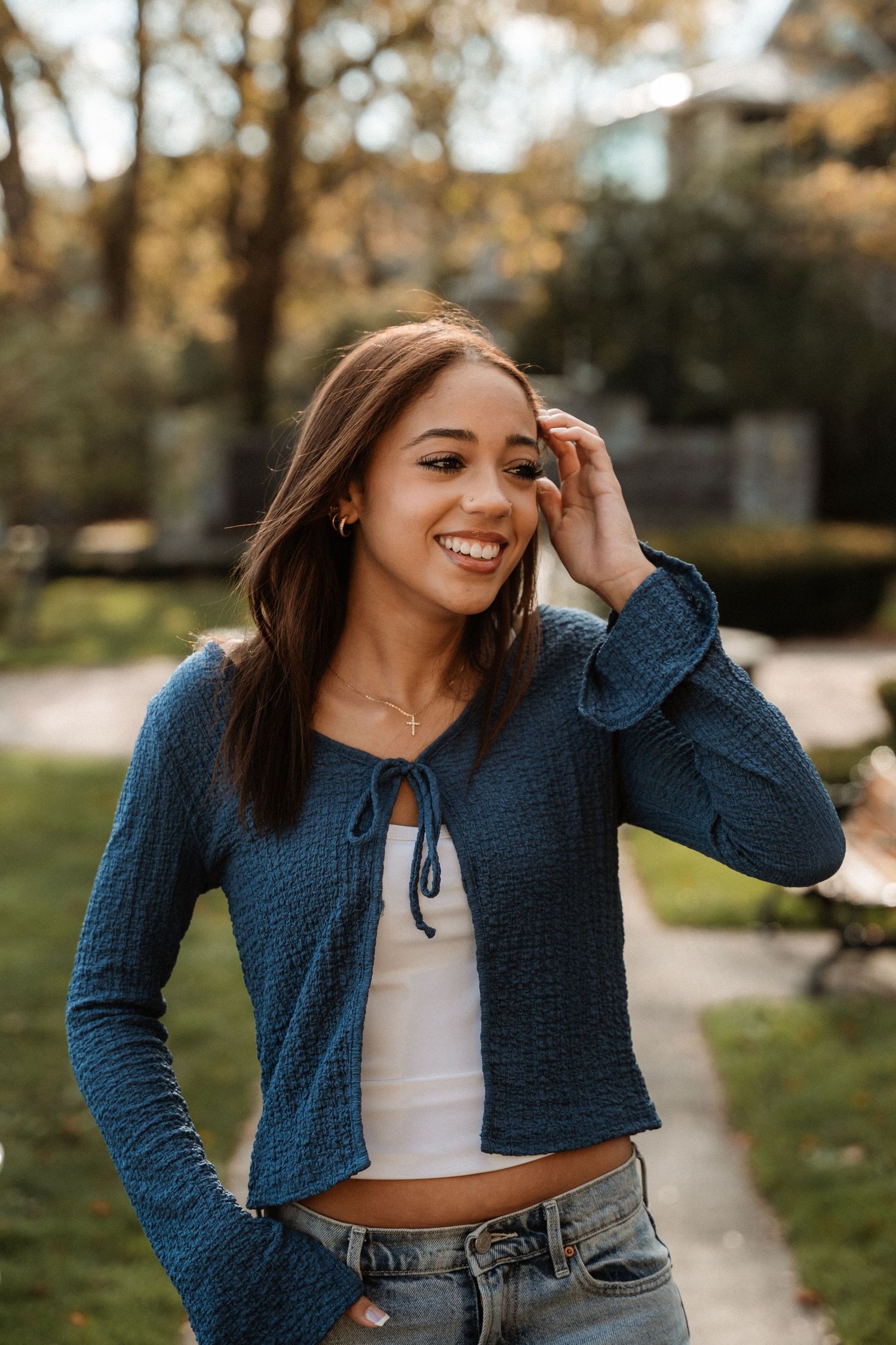 Smiling young woman in blue cardigan and jeans poses outdoors in a park during autumn.