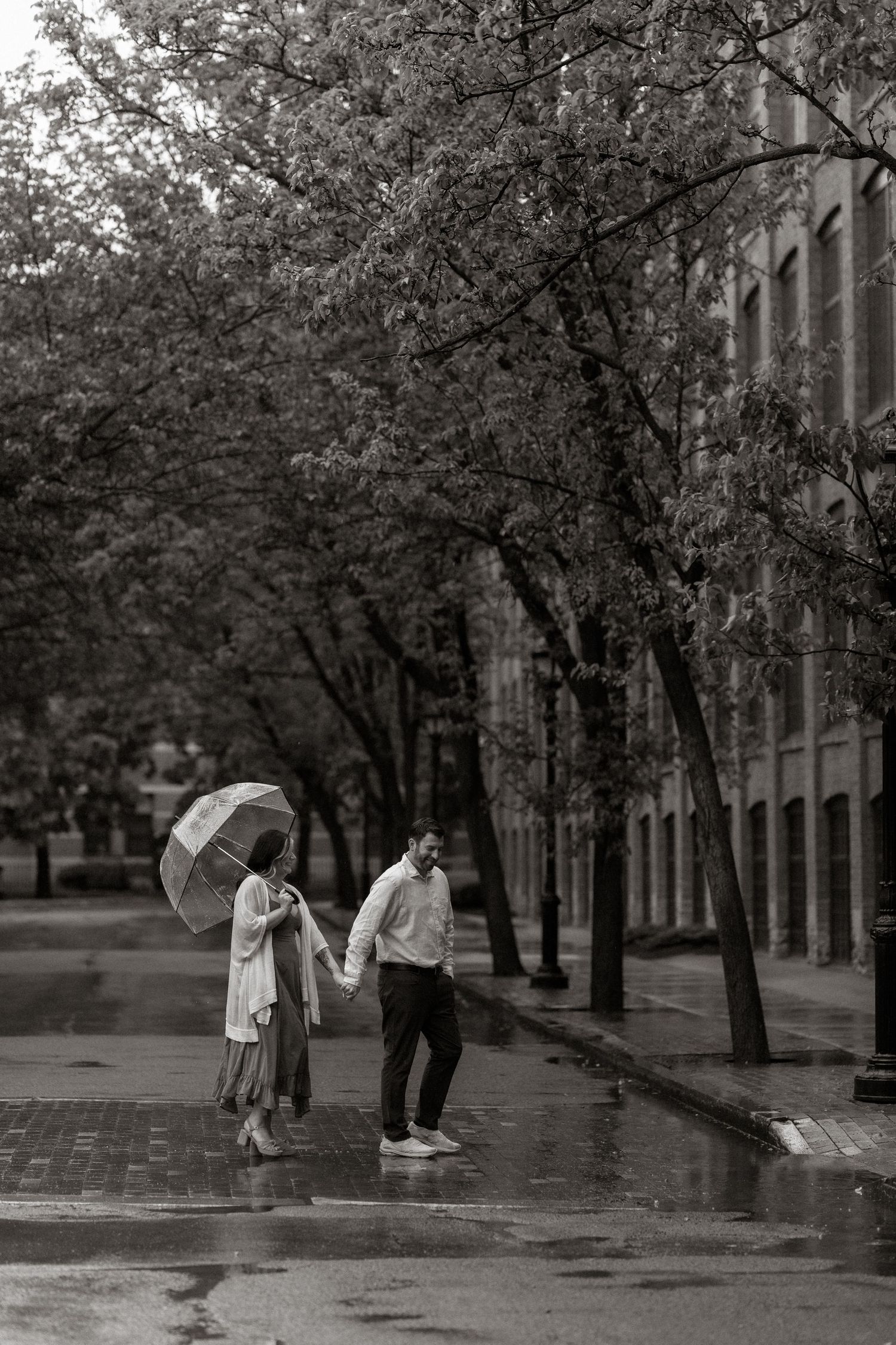 Black and white photo of a couple walking hand in hand in the rain under a clear umbrella on a wet city street.
