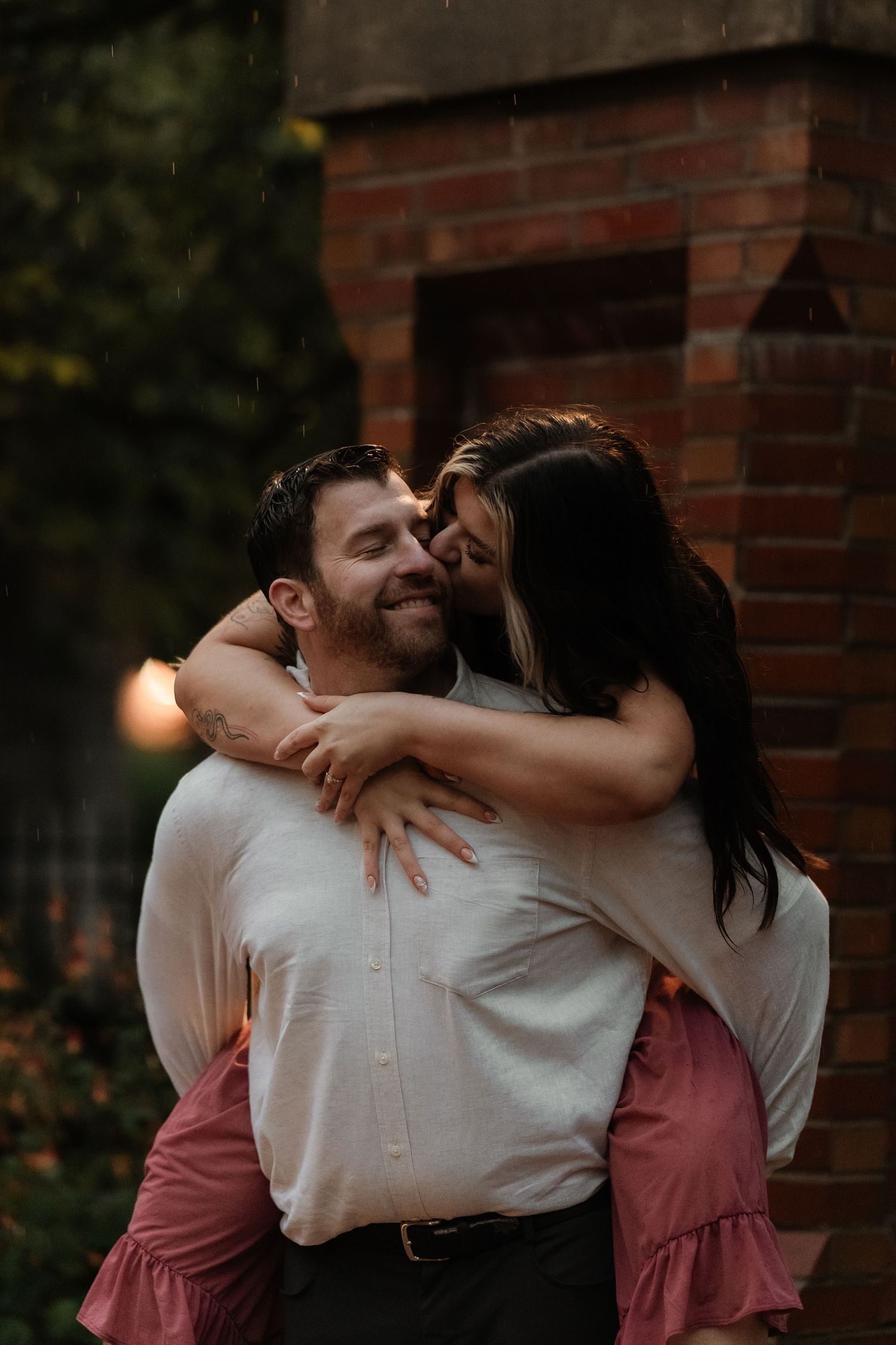 A smiling man gives a woman a piggyback ride as she kisses his cheek near a brick wall.