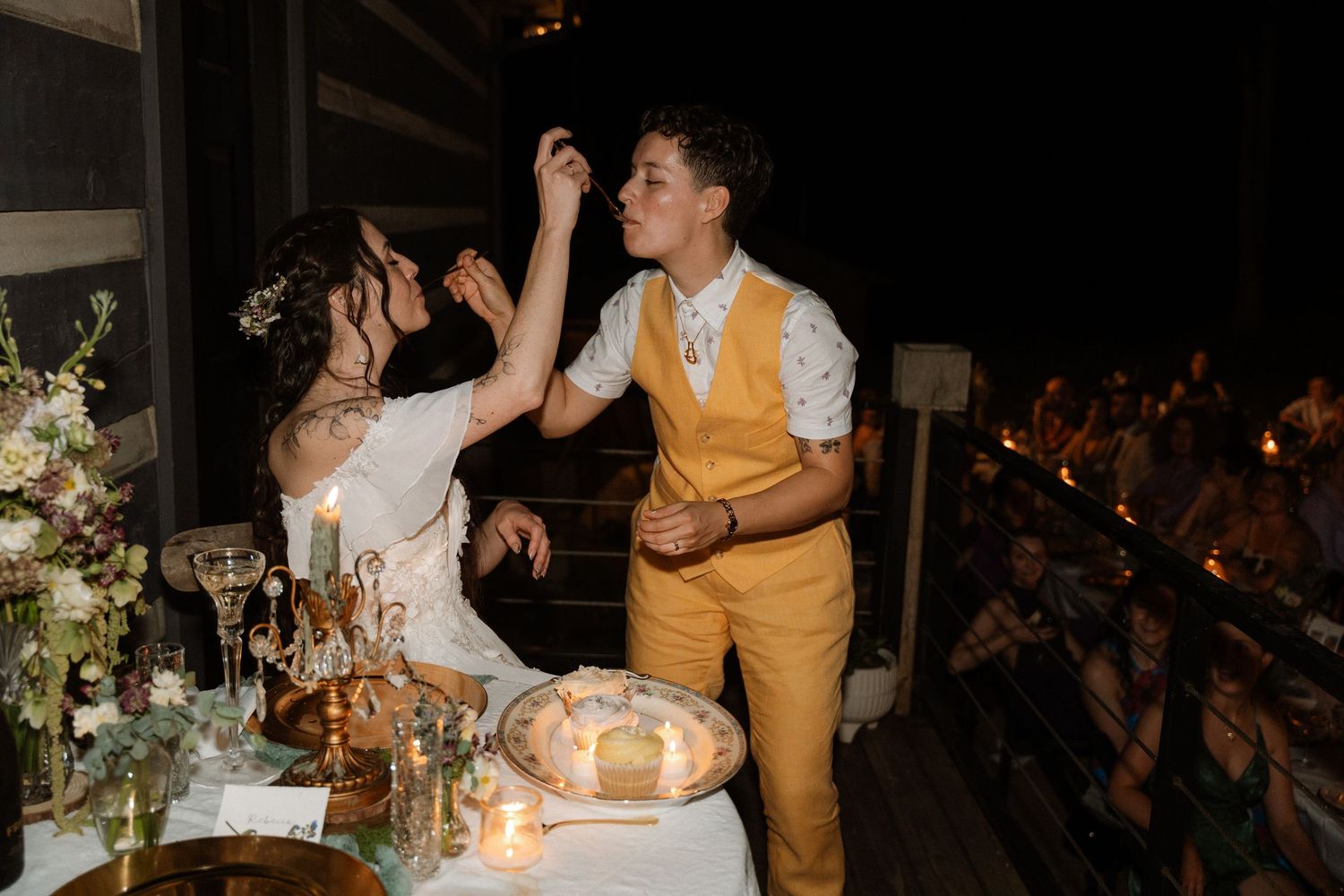 Bride and groom sharing wedding cake at outdoor reception, groom in yellow vest feeding cake to bride at candlelit table.