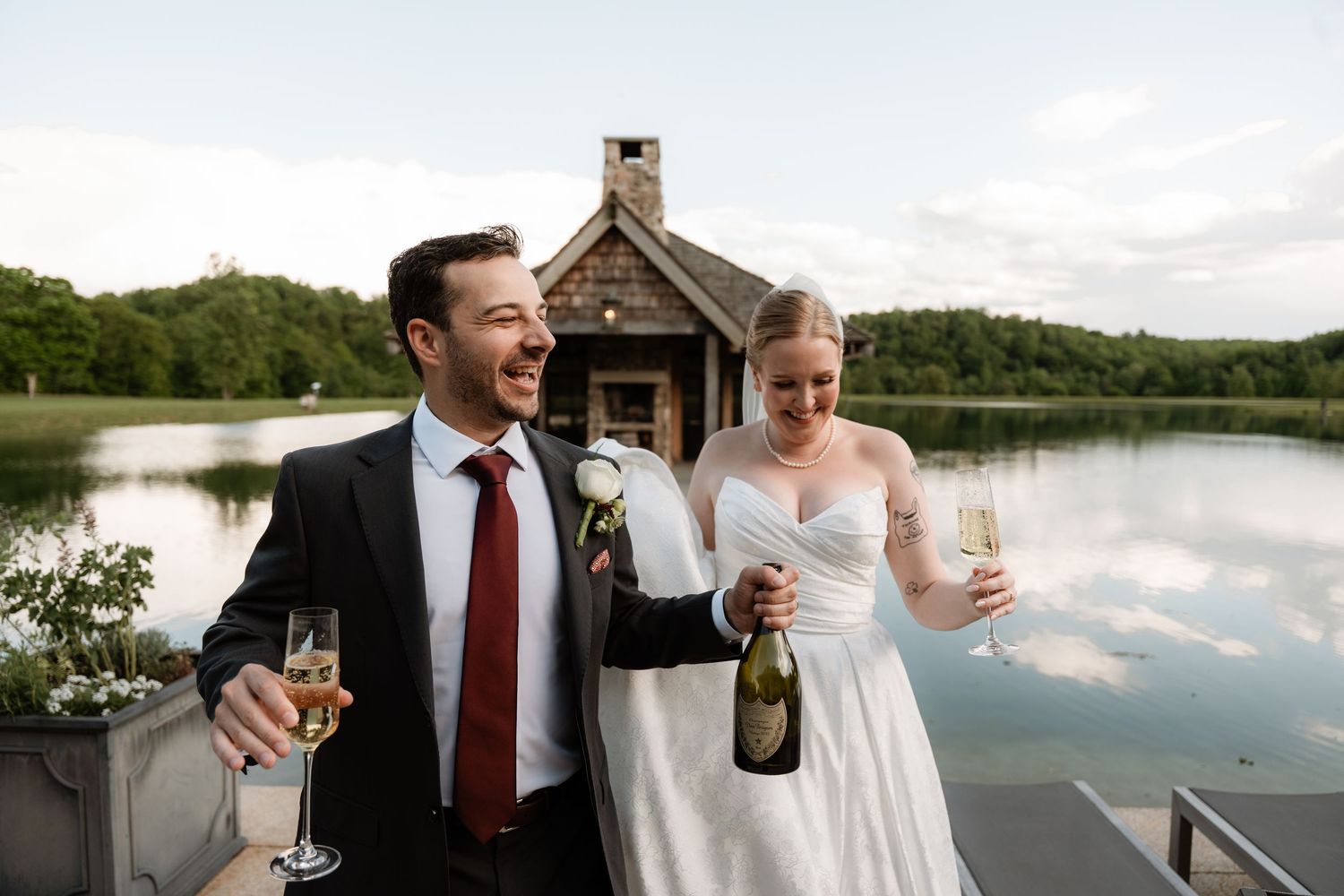 Bride and groom celebrating with champagne by a lakeside chapel on their wedding day.