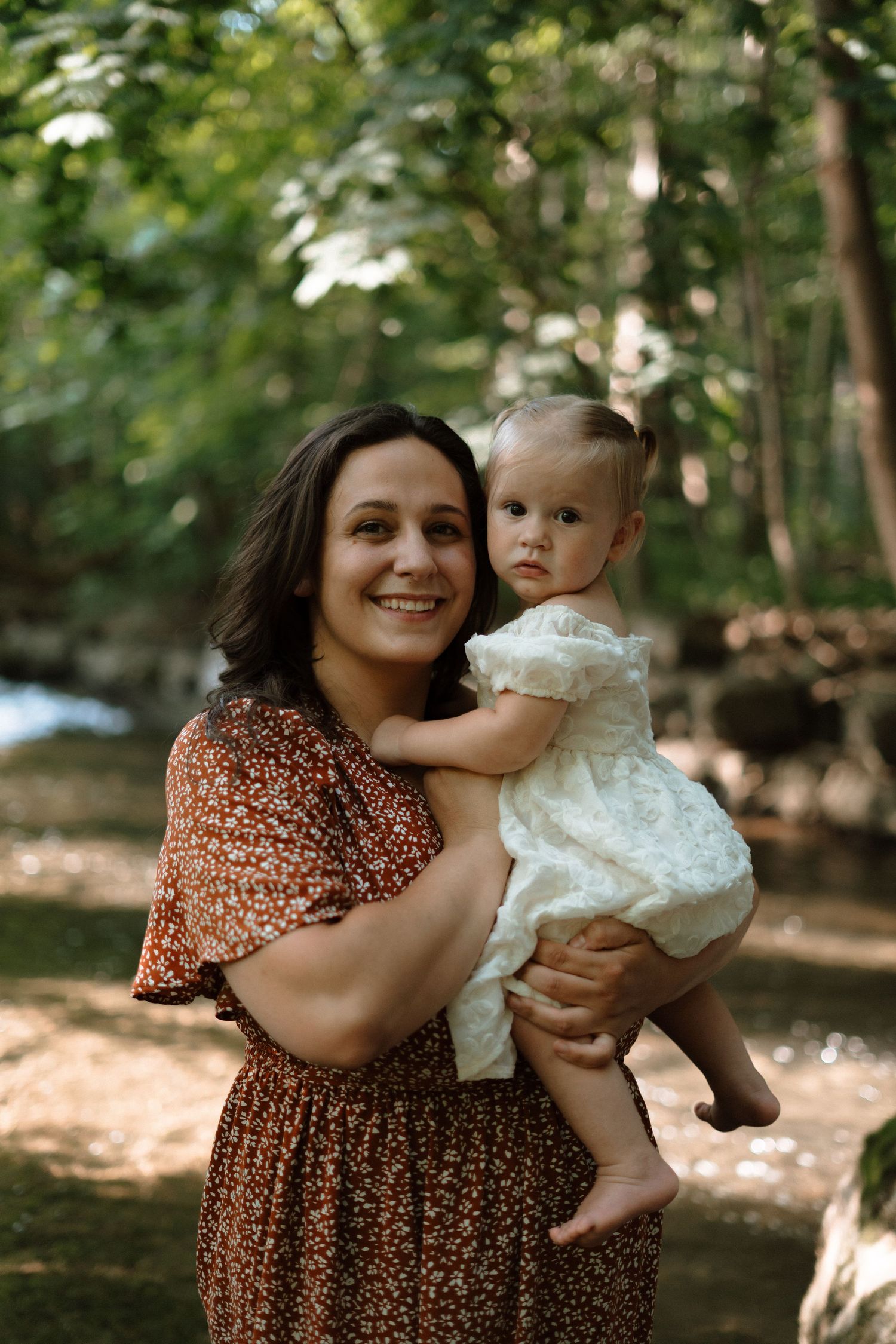 A smiling woman in a floral dress holds a baby girl in a white dress outdoors in a lush green forest setting.