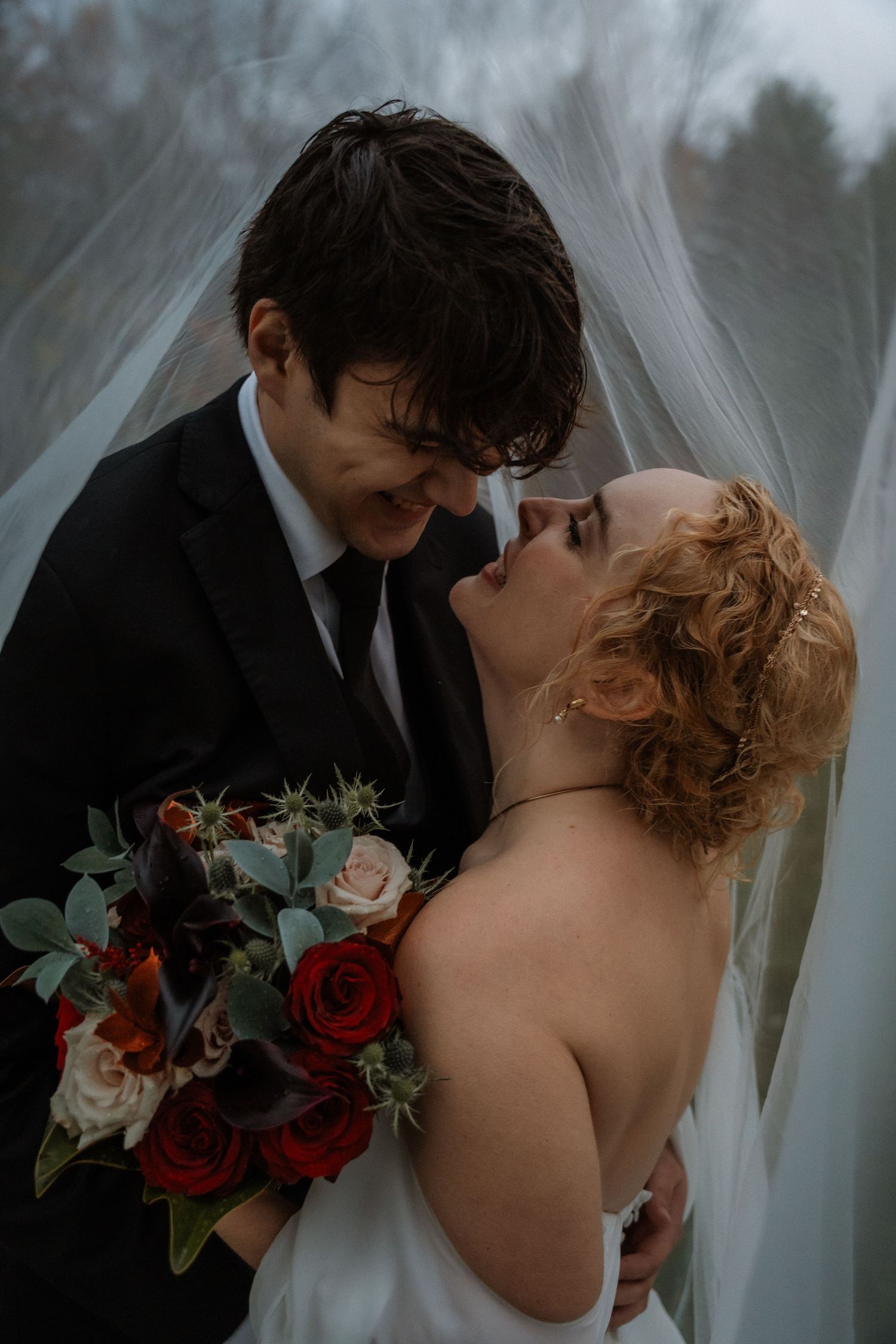 A romantic wedding couple shares an intimate kiss behind sheer white curtains with red and pink roses in their hands.