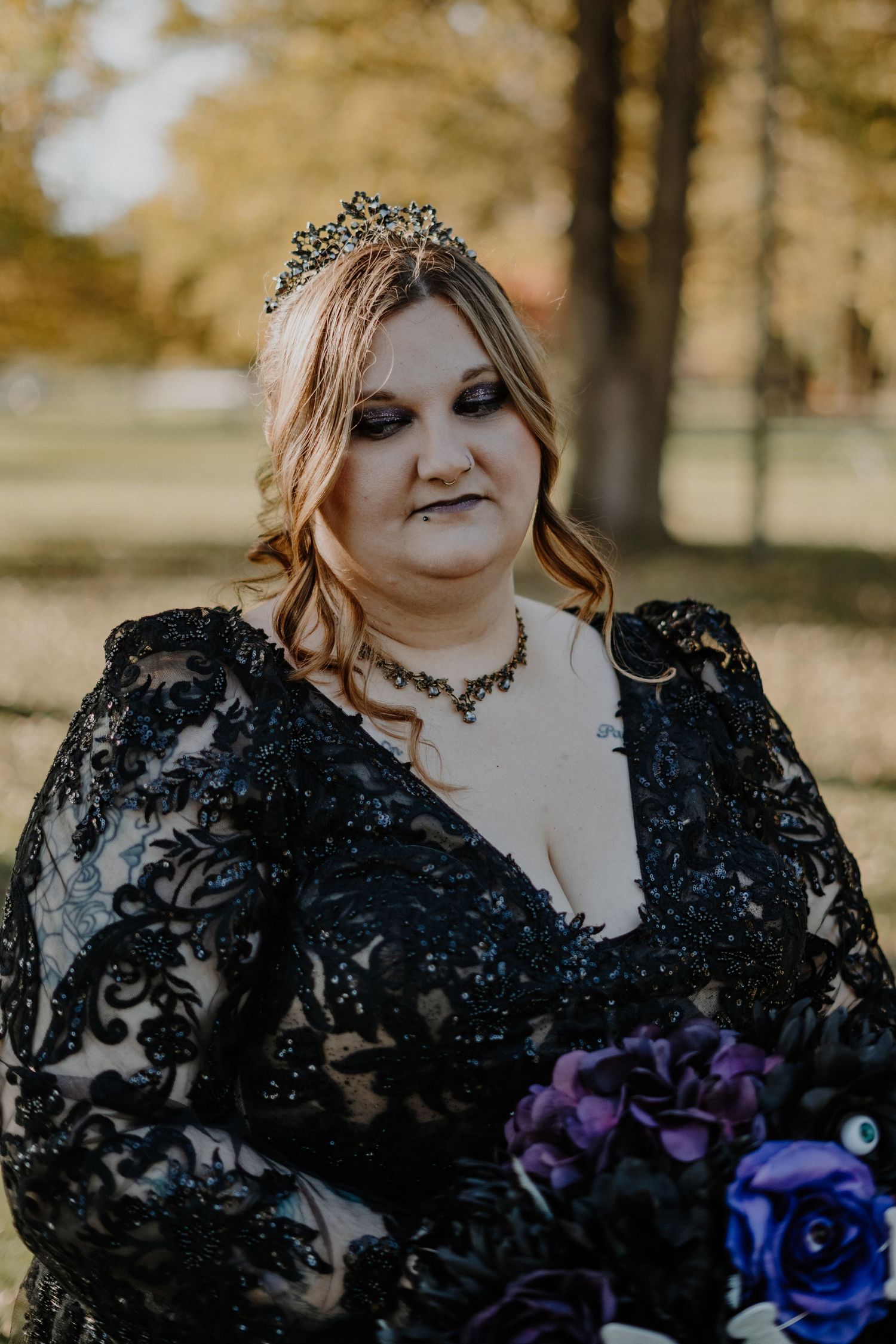 Bride in black lace wedding gown poses with purple bouquet in autumn outdoor setting.