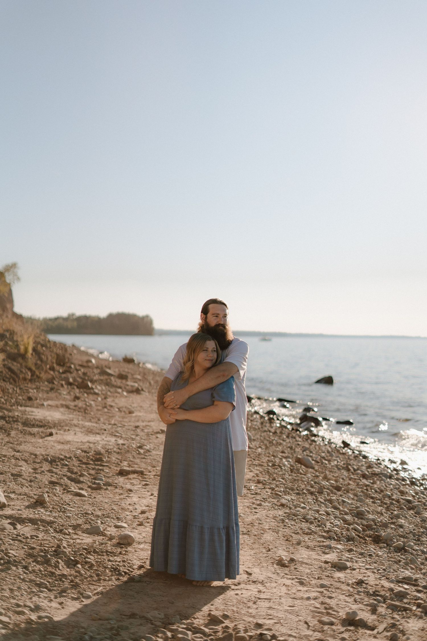 A couple embraces on a rocky beach shoreline at sunset with calm waters and cliffs in the background.