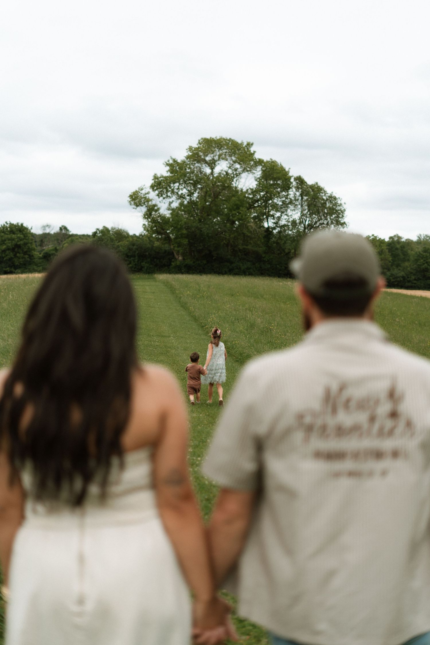 Two people stand together watching someone in the distance on a grassy green hill under an overcast sky.