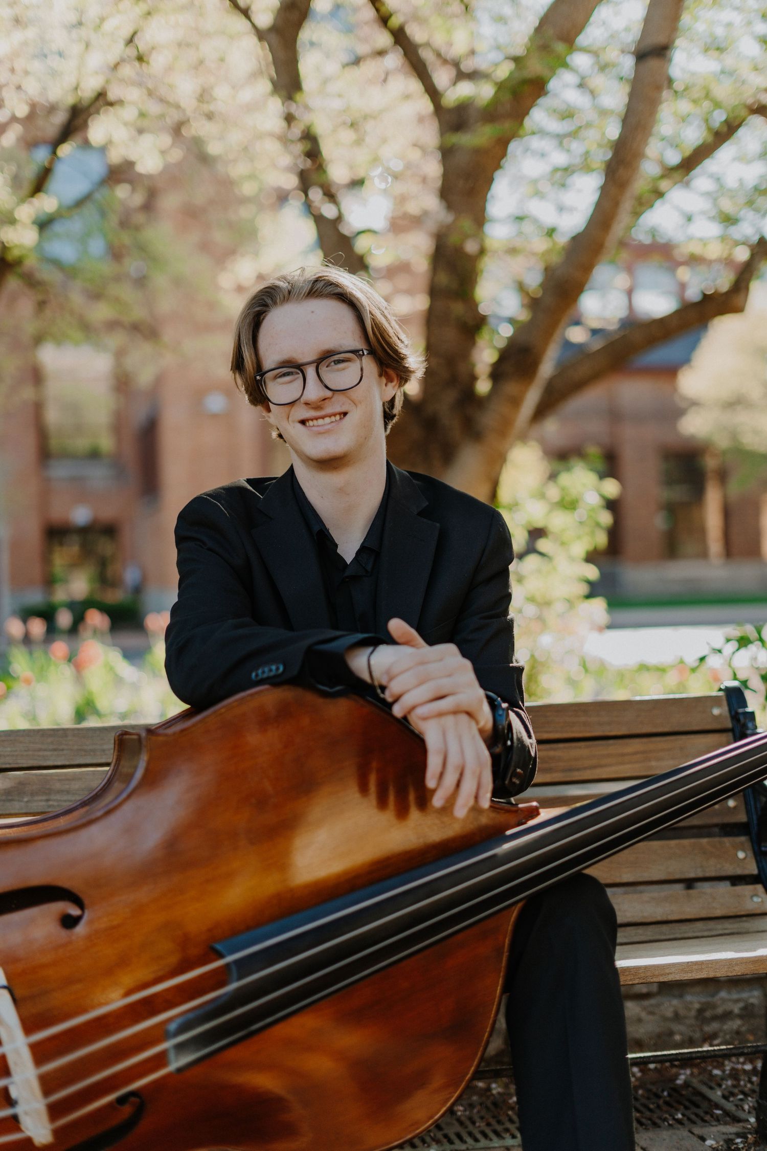 Senior portrait where person sits on a bench with a bass during cherry blossom season.