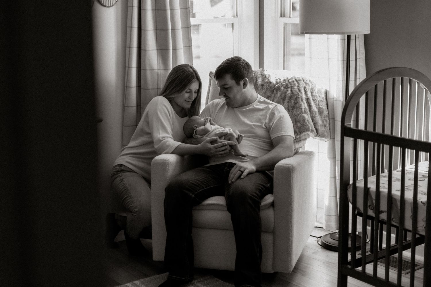 Black and white photo of new parents sitting in armchair together, lovingly gazing at their newborn baby.