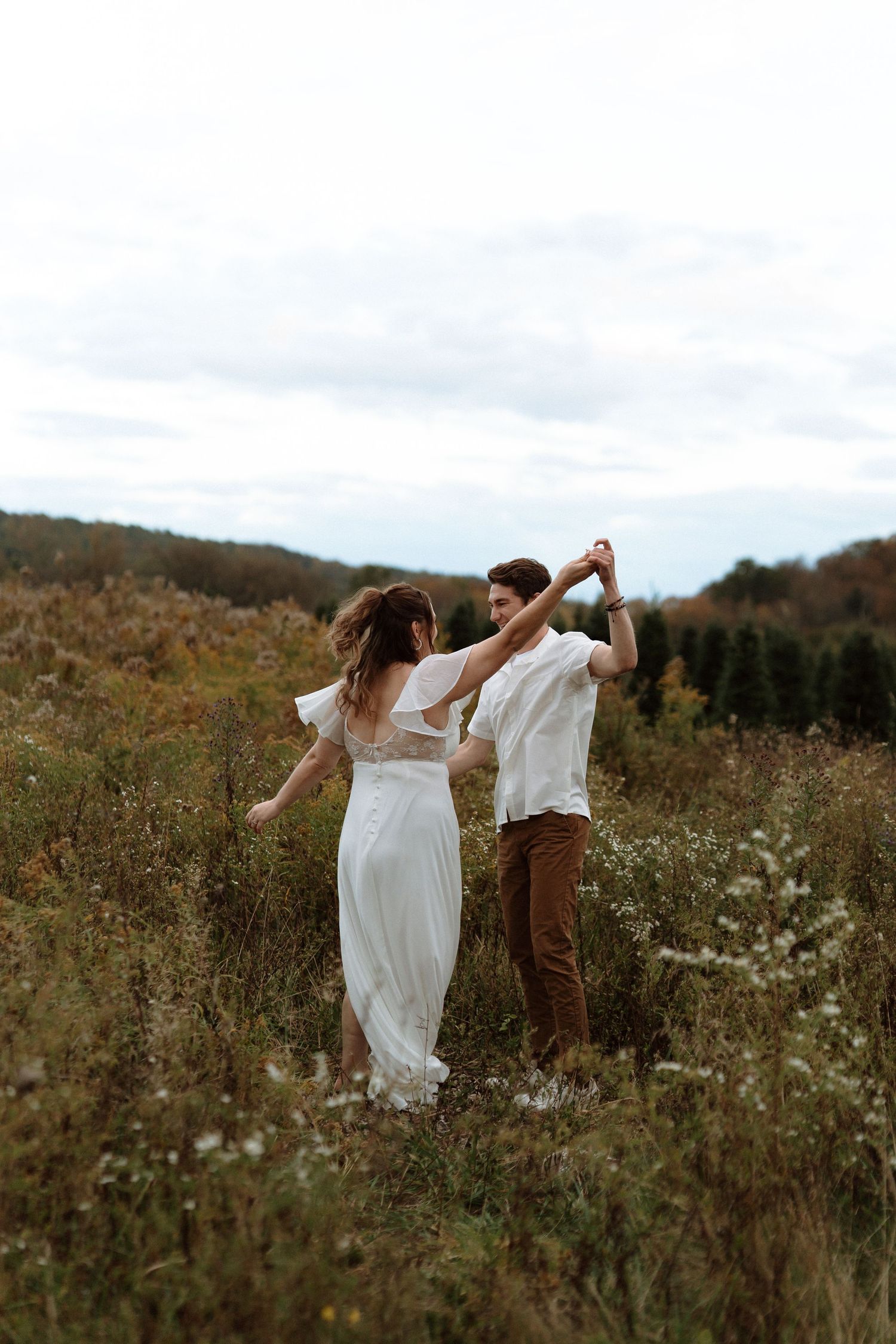 A couple dances together in a wildflower meadow wearing white and brown outfits at sunset.