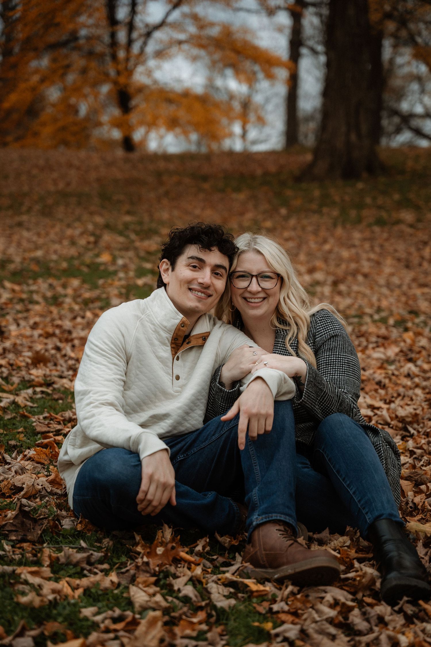 Couple sitting together on autumn leaves in a park, smiling, wearing cozy sweaters and jeans during fall season.