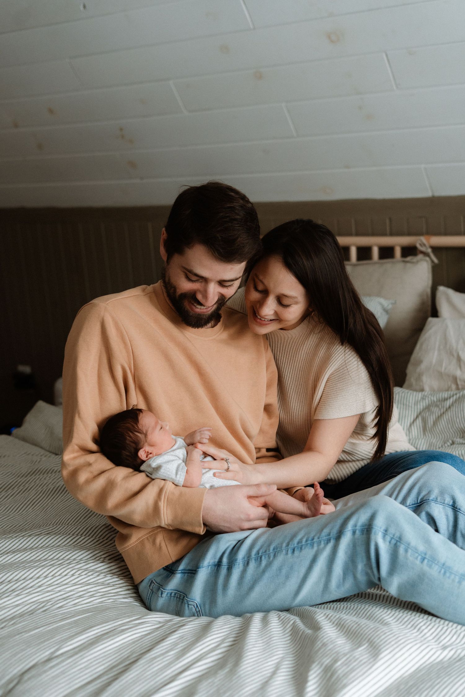 Happy young parents sitting on bed, smiling at their newborn baby, in a cozy bedroom setting.