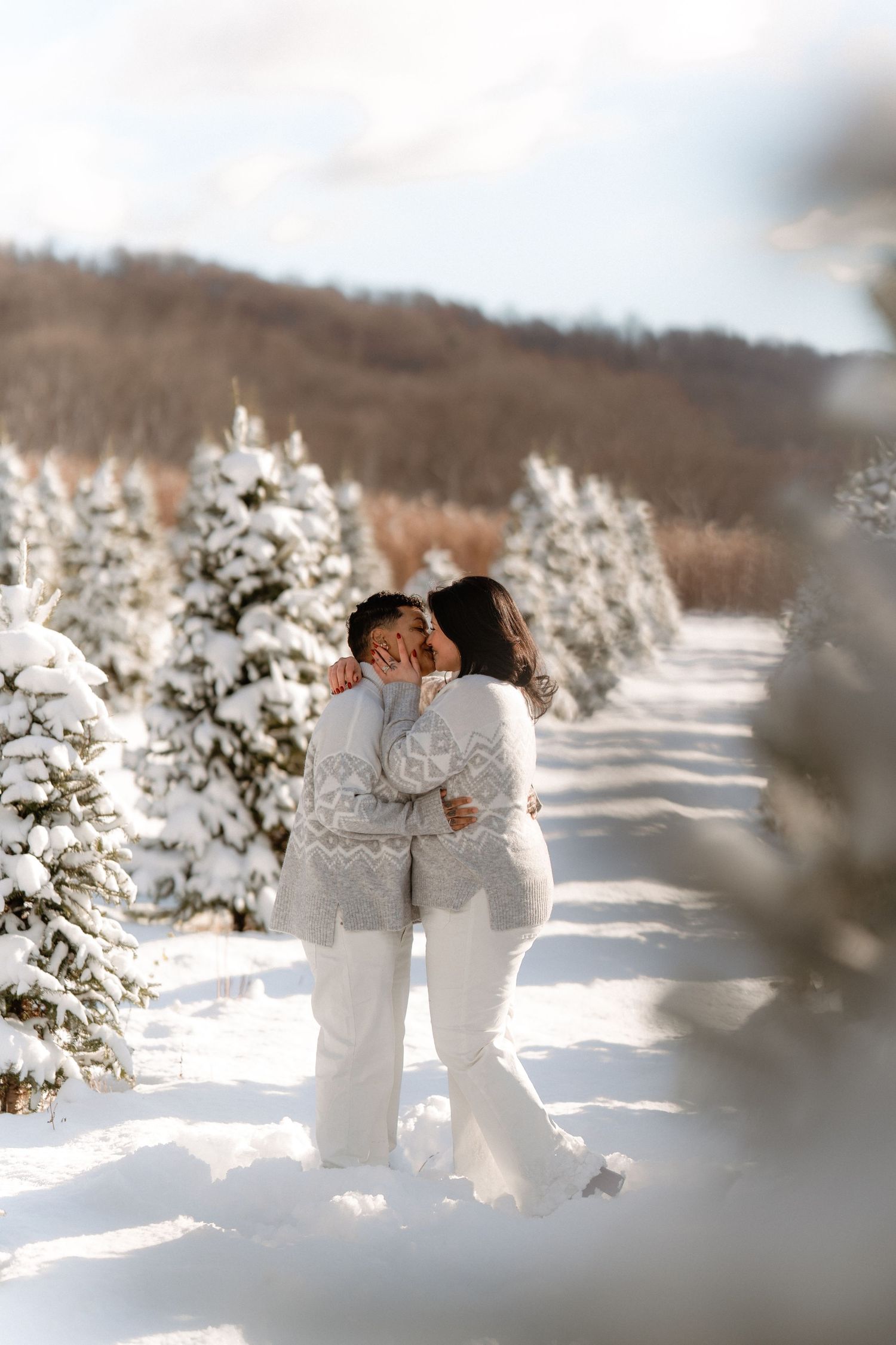 Couple kissing in snowy winter forest surrounded by snow-covered evergreen trees wearing matching white Nordic sweaters.