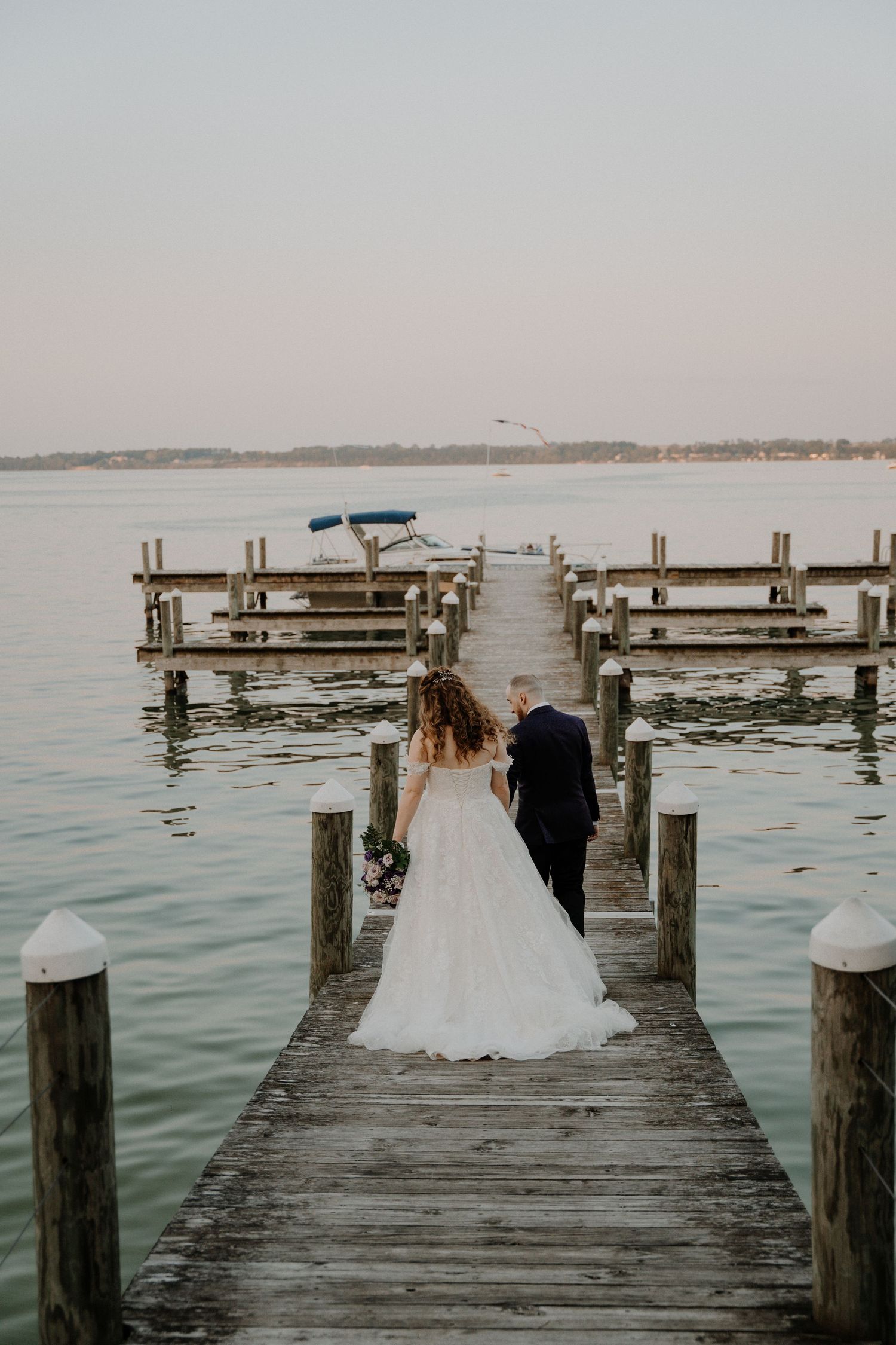 Wedding couple walking on a wooden pier overlooking a serene lake at sunset.