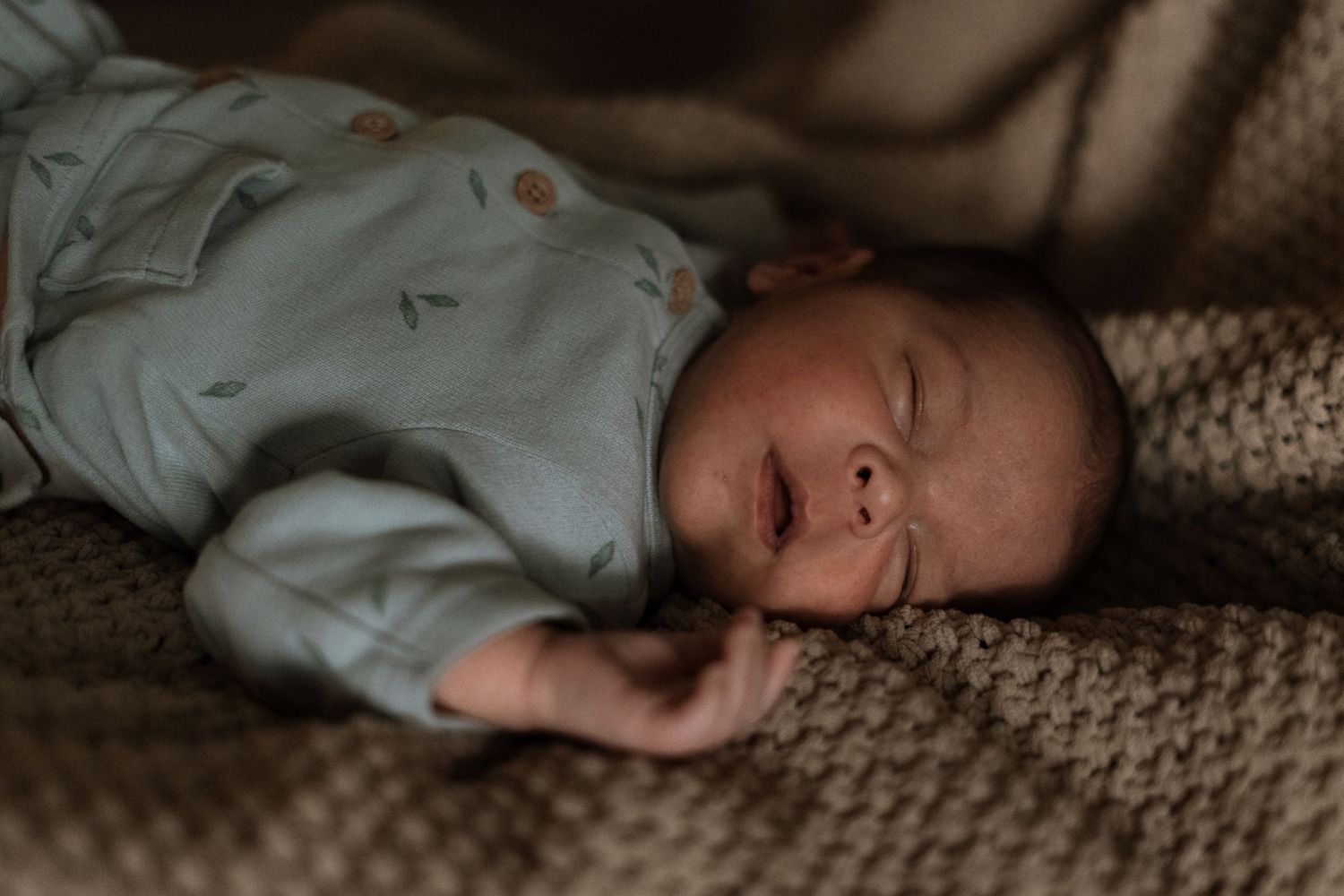 Baby wearing light blue onesie lying on textured knit blanket in moody lighting.