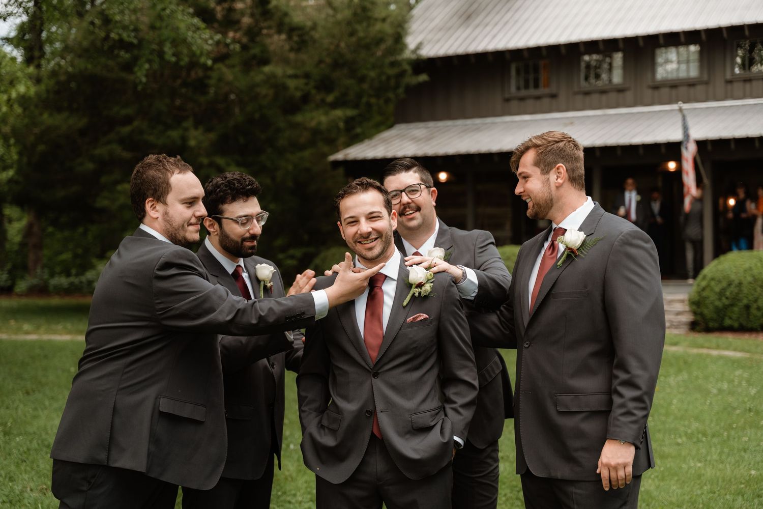 Groom surrounded by smiling groomsmen in gray suits with burgundy ties, sharing a joyful moment outdoors near a rustic barn.