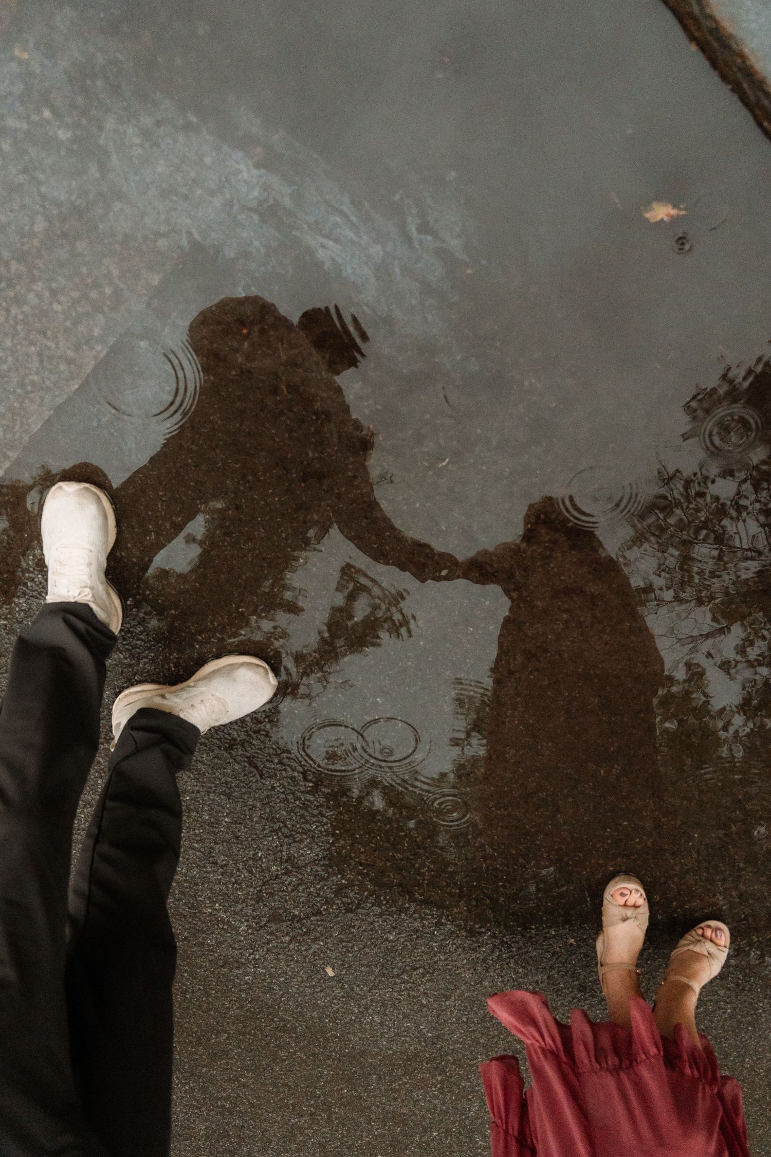 Two people holding hands reflected in a rain puddle, one wearing white boots and the other in a red skirt.