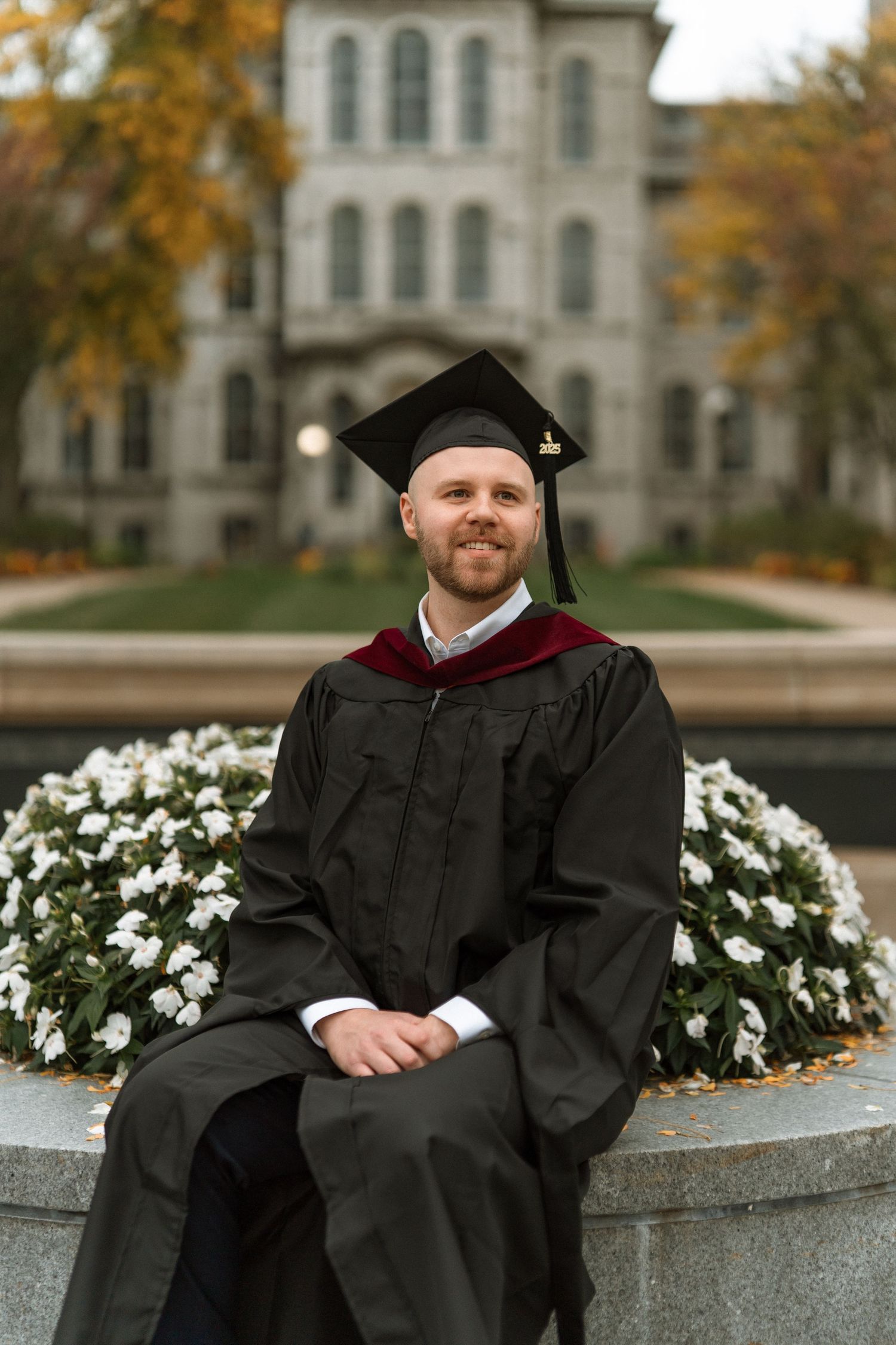 Graduate in cap and gown sits on stone ledge with white flowers and autumn foliage in background.