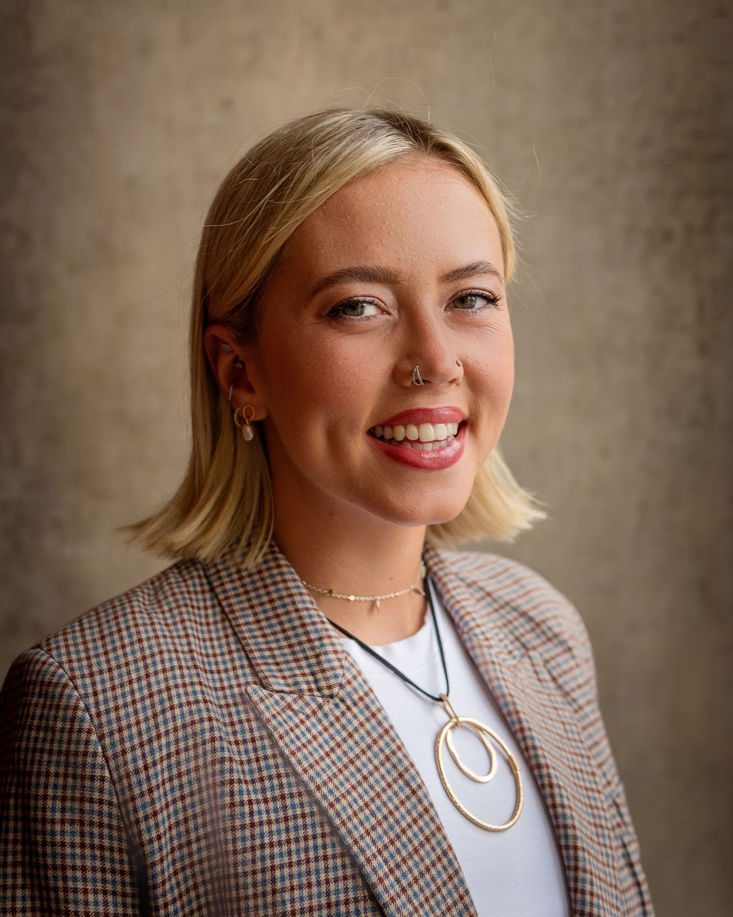 Professional portrait of someone wearing a plaid blazer, white top and circular necklace with blonde hair and warm smile.