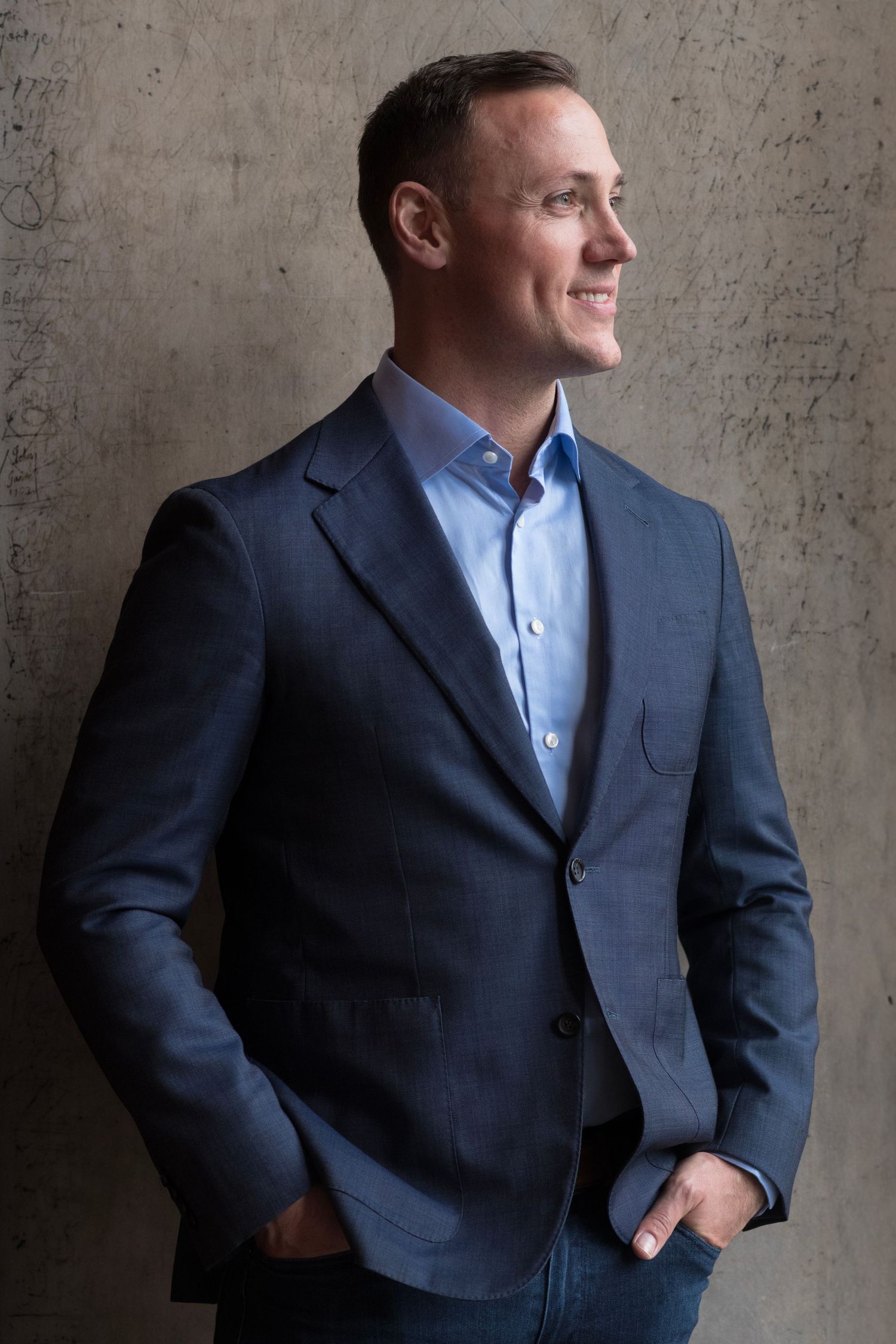 Professional looking sideways while leaning against textured wall wearing navy suit jacket and light blue shirt.
