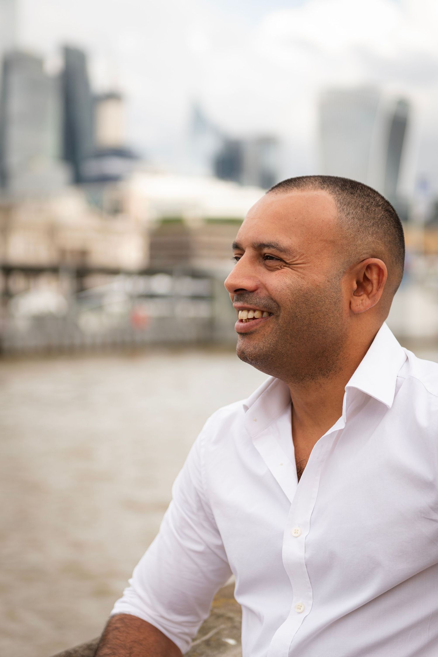 Man in white shirt smiling while sitting outdoors with city skyline in background.