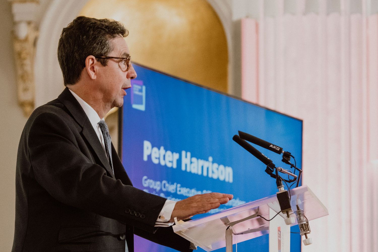 Speaker presenting at a podium with Peter Harrison text visible on blue background.