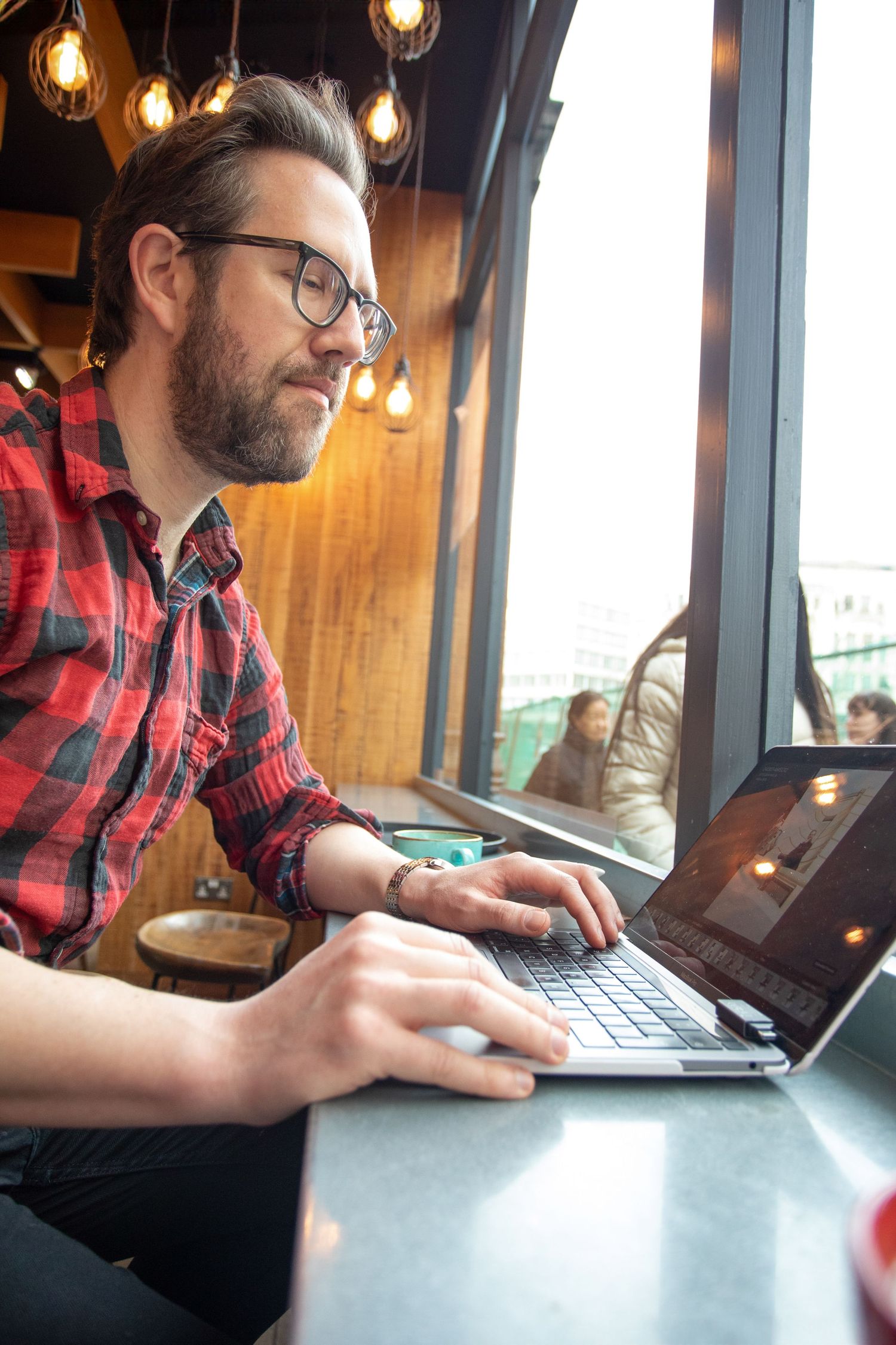 Man in plaid shirt working on laptop by window in coffee shop with pendant lights.