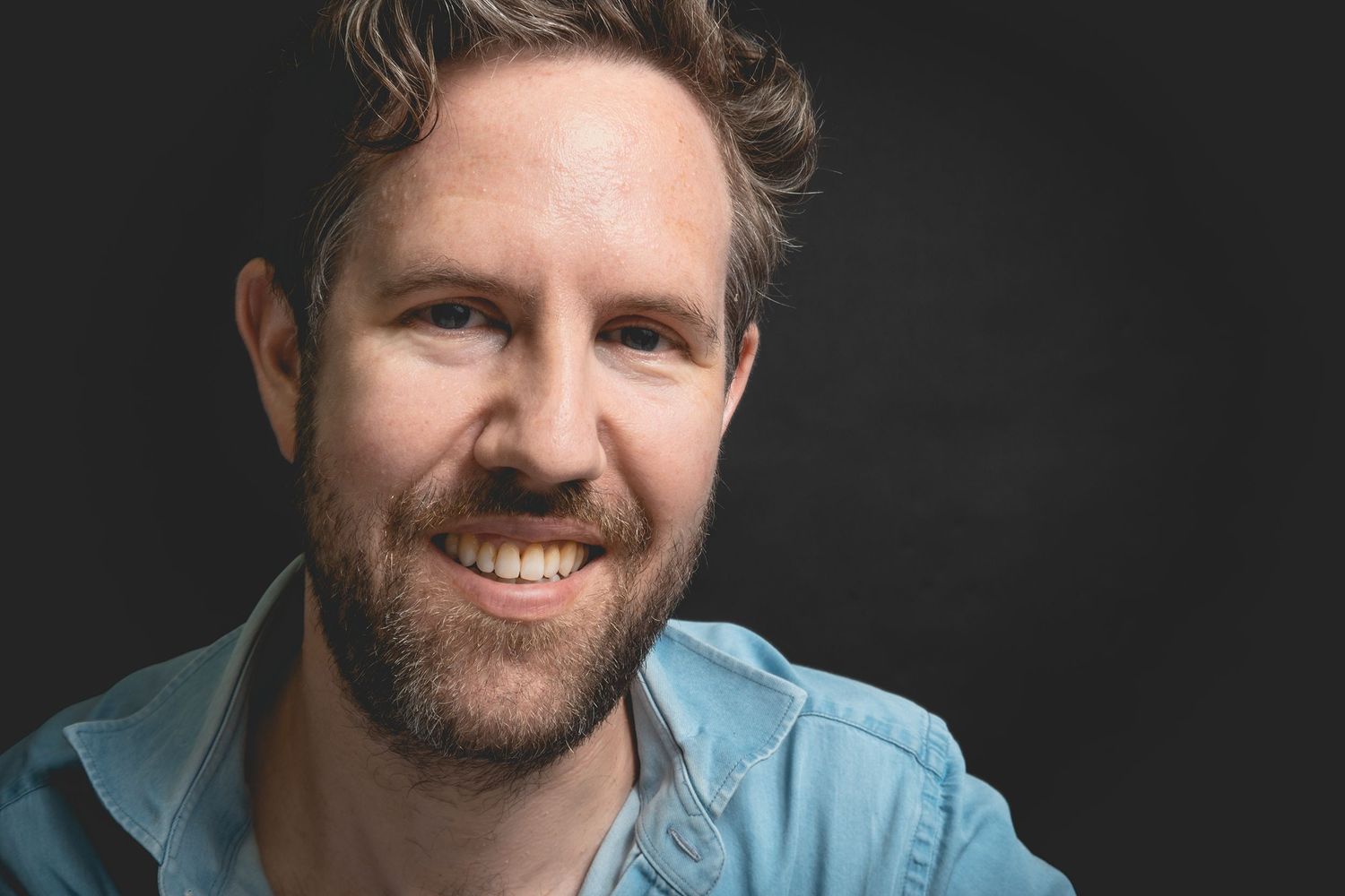Bearded man wearing light blue button down shirt smiles warmly against dark background in studio portrait.