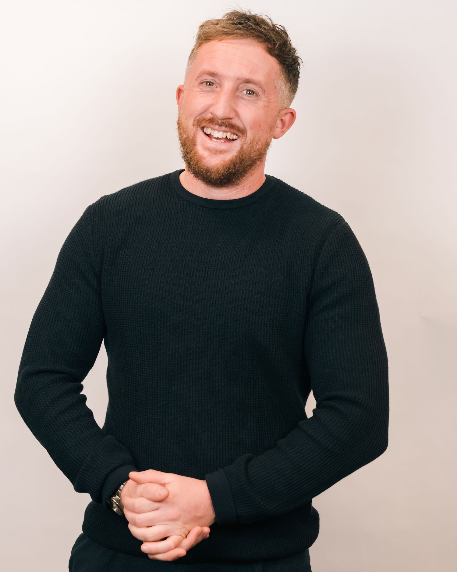 Candid portrait of person in black sweater smiling warmly against white studio background.