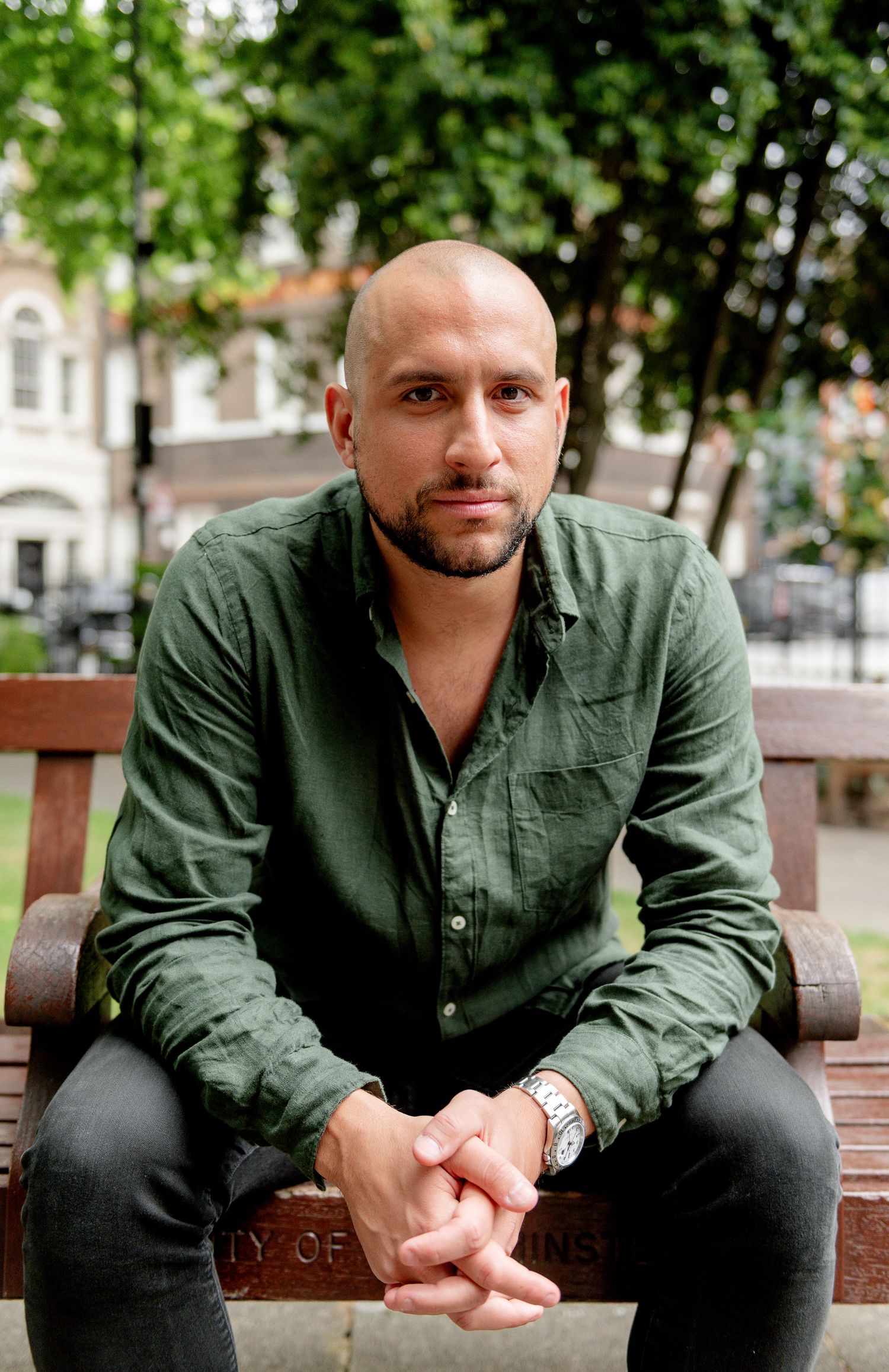 A person in a green button-down shirt sits on a wooden bench outdoors in an urban setting.