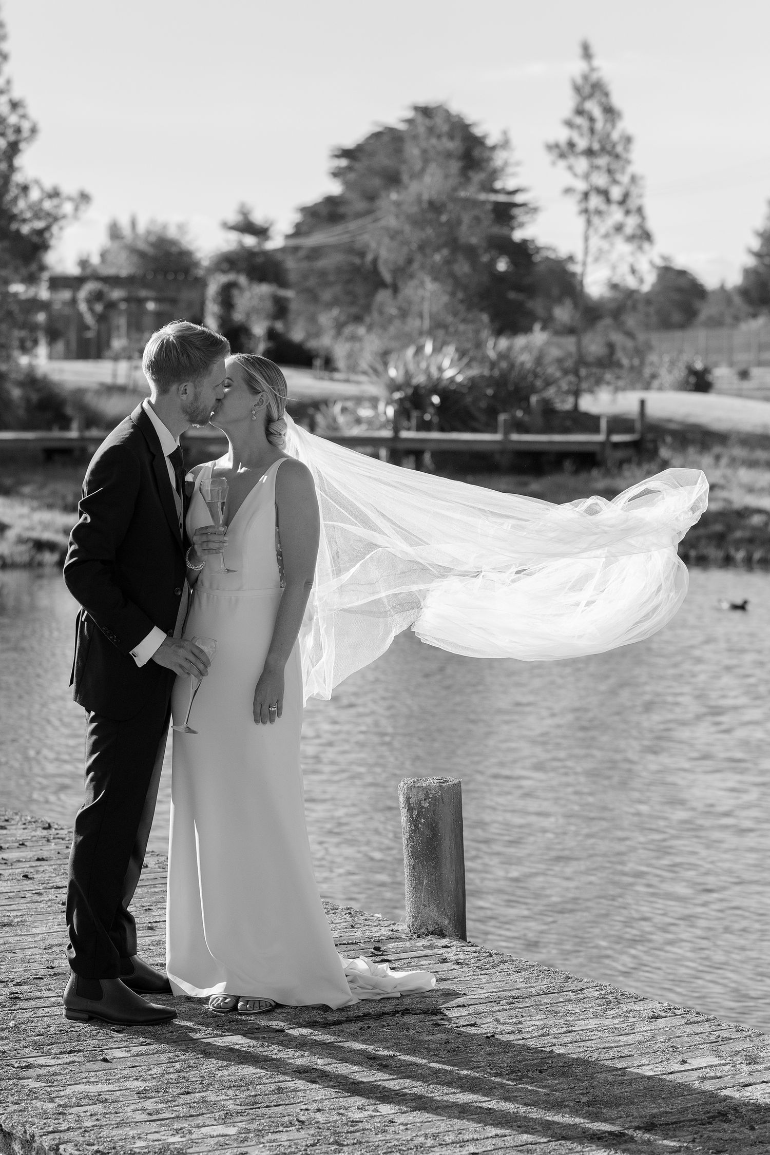 Newlyweds embrace by waterfront as bride's veil flows dramatically in black and white photo.