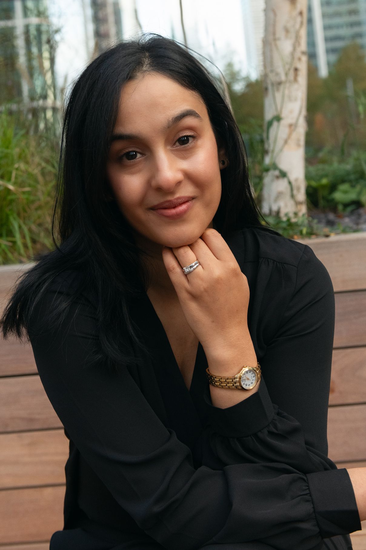 Smiling woman in black blouse poses outdoors while showing jewelry and watch accessories.