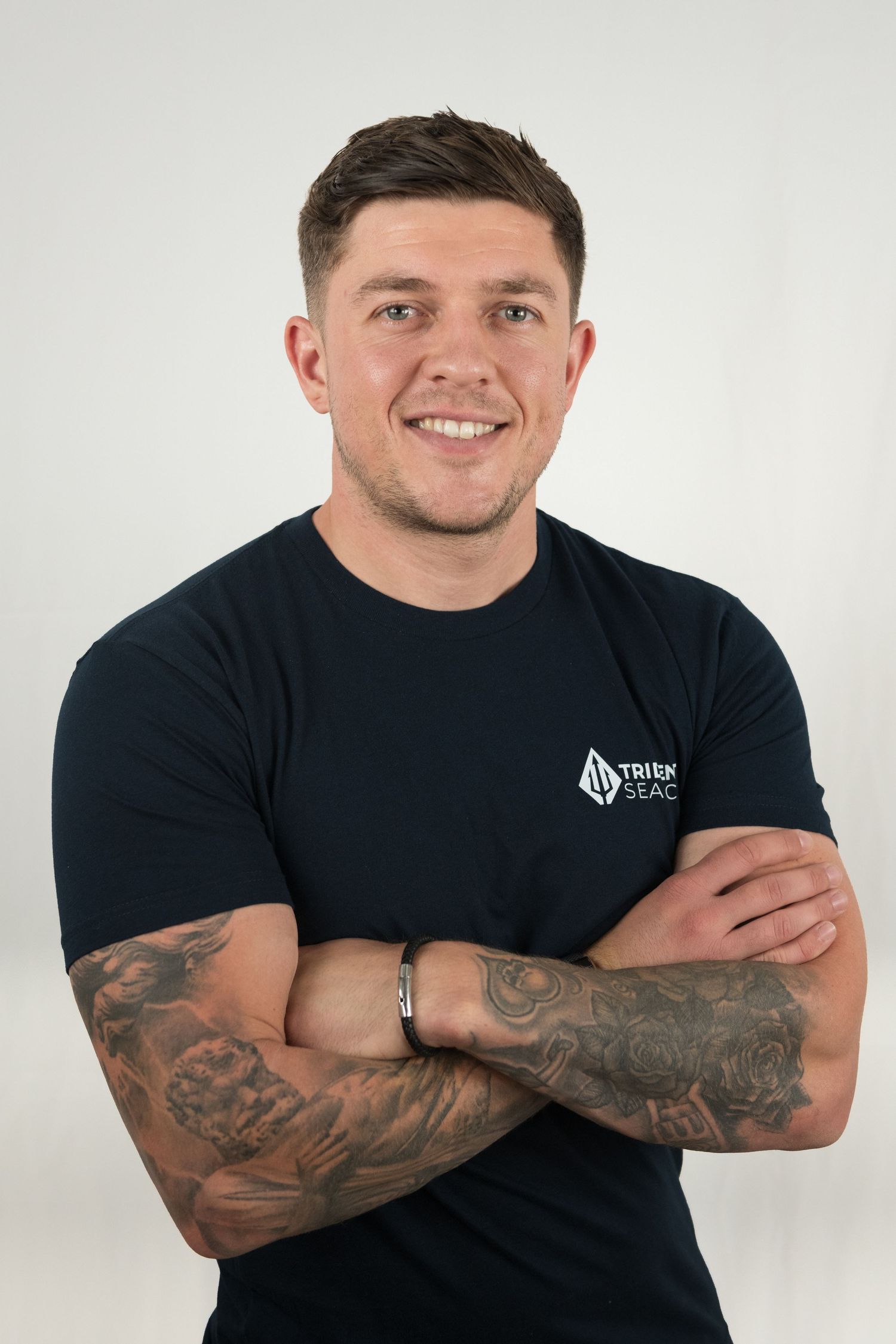 Team member in black Trident Search t-shirt with tattooed arms crossed poses against white background.