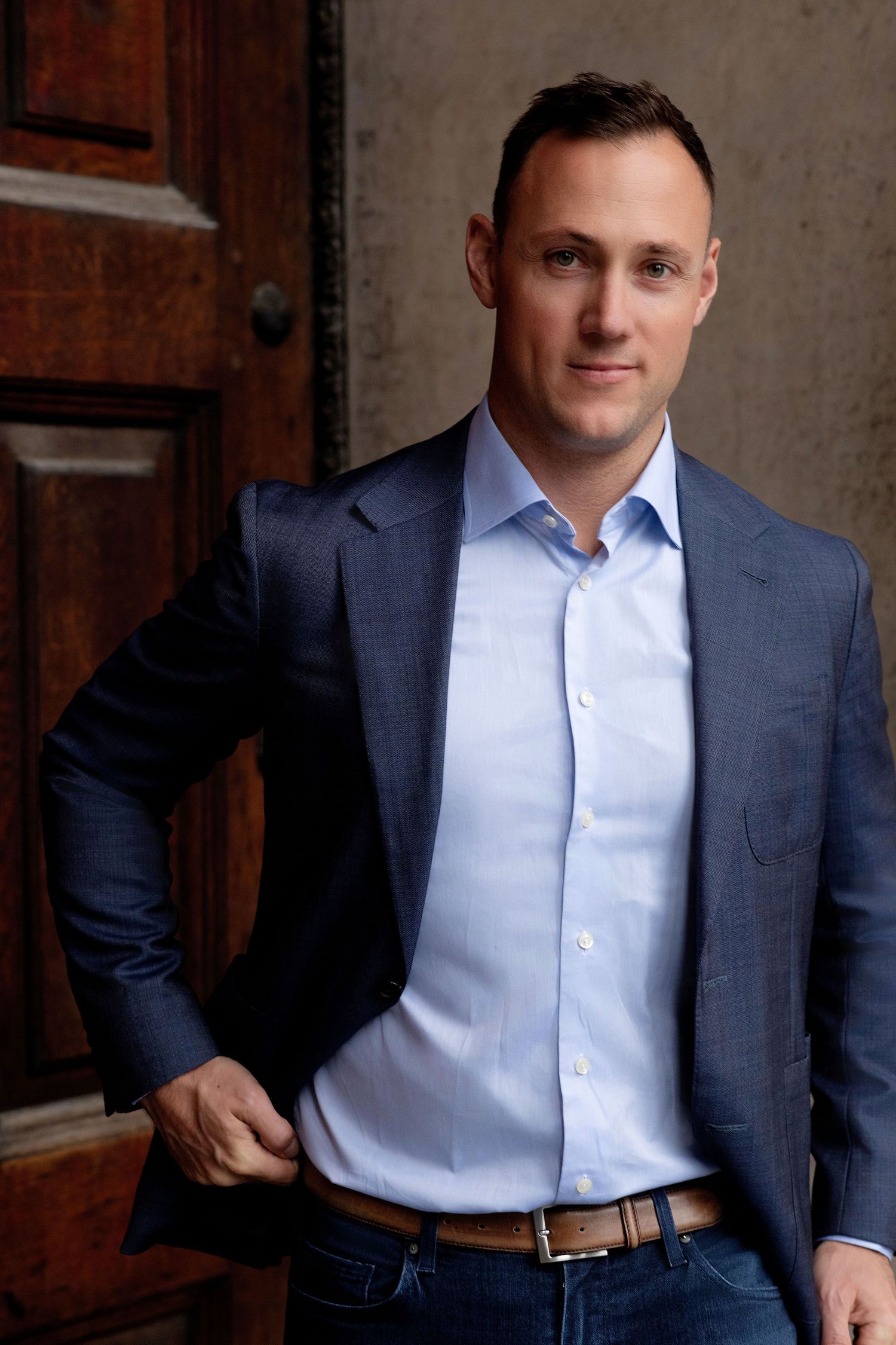 Professional headshot portrait of someone in a navy suit jacket and light blue dress shirt against dark background.