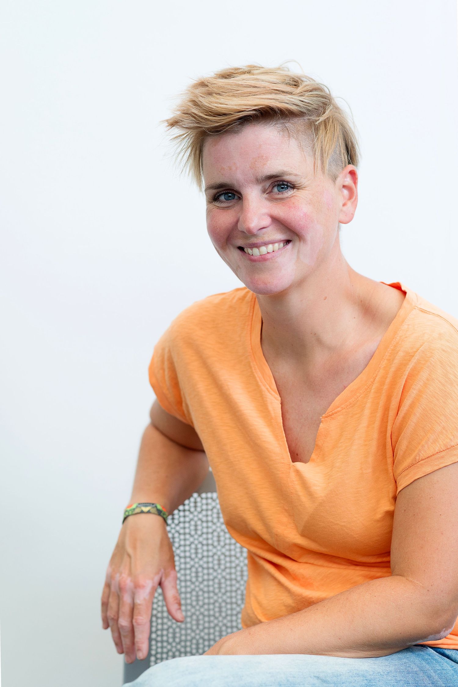 Person with short blonde hair wearing an orange t-shirt sits casually and smiles against a light background.