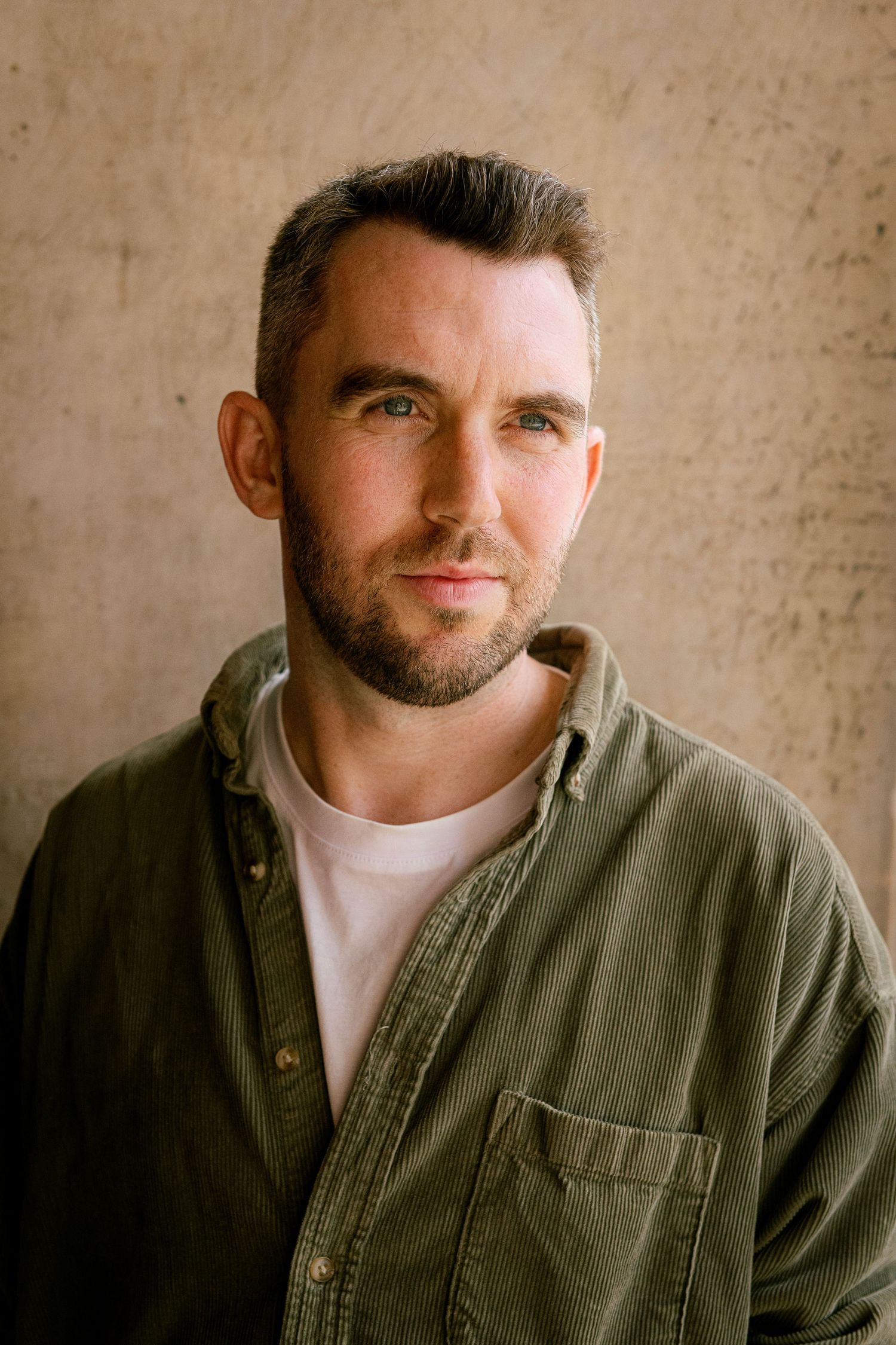Close-up portrait of someone wearing an olive green button-down shirt over a white t-shirt against a textured beige wall.