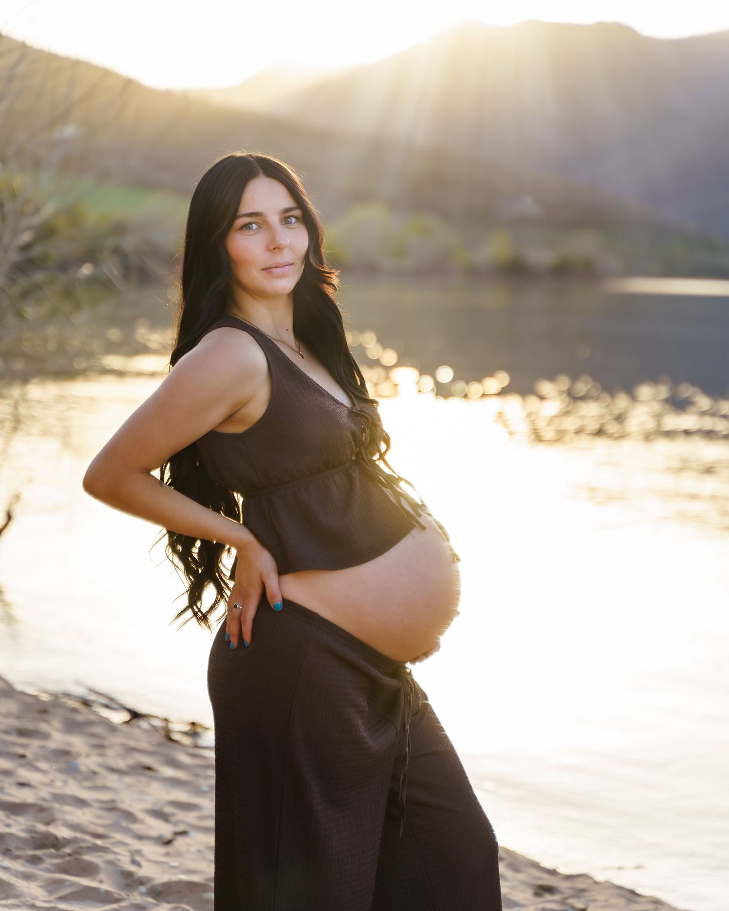 Pregnant woman in black dress poses on sandy beach at golden hour with mountains and lake behind her.
