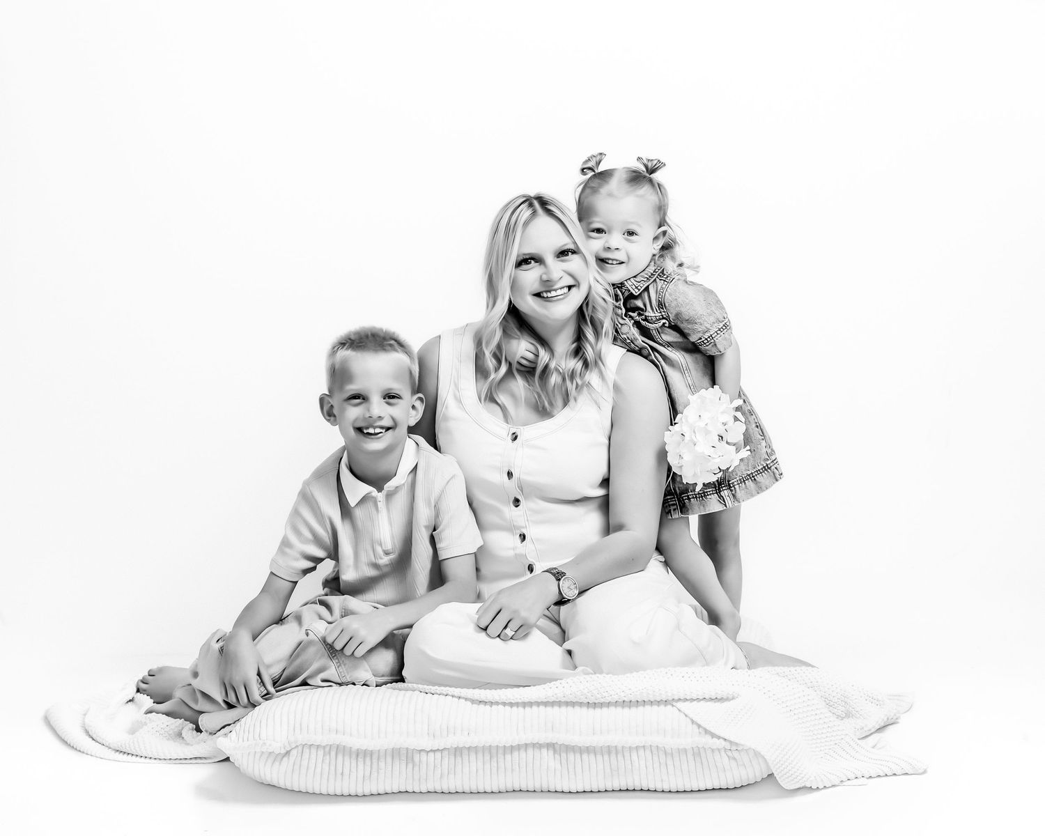 Black and white studio portrait of smiling blonde mother sitting with young son and toddler daughter.