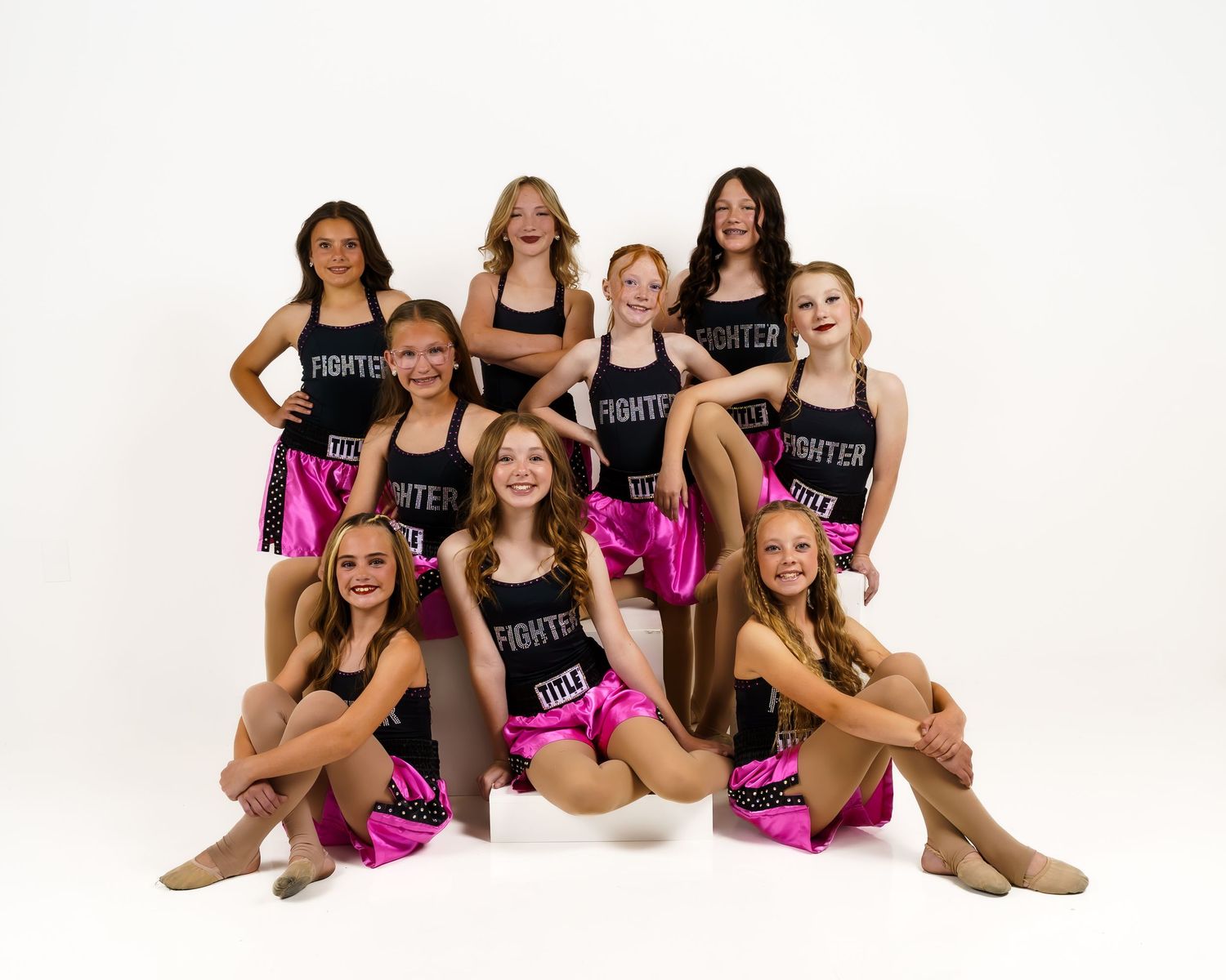 Nine young dancers in matching black and pink Fighter costumes pose together for a group dance recital photo.
