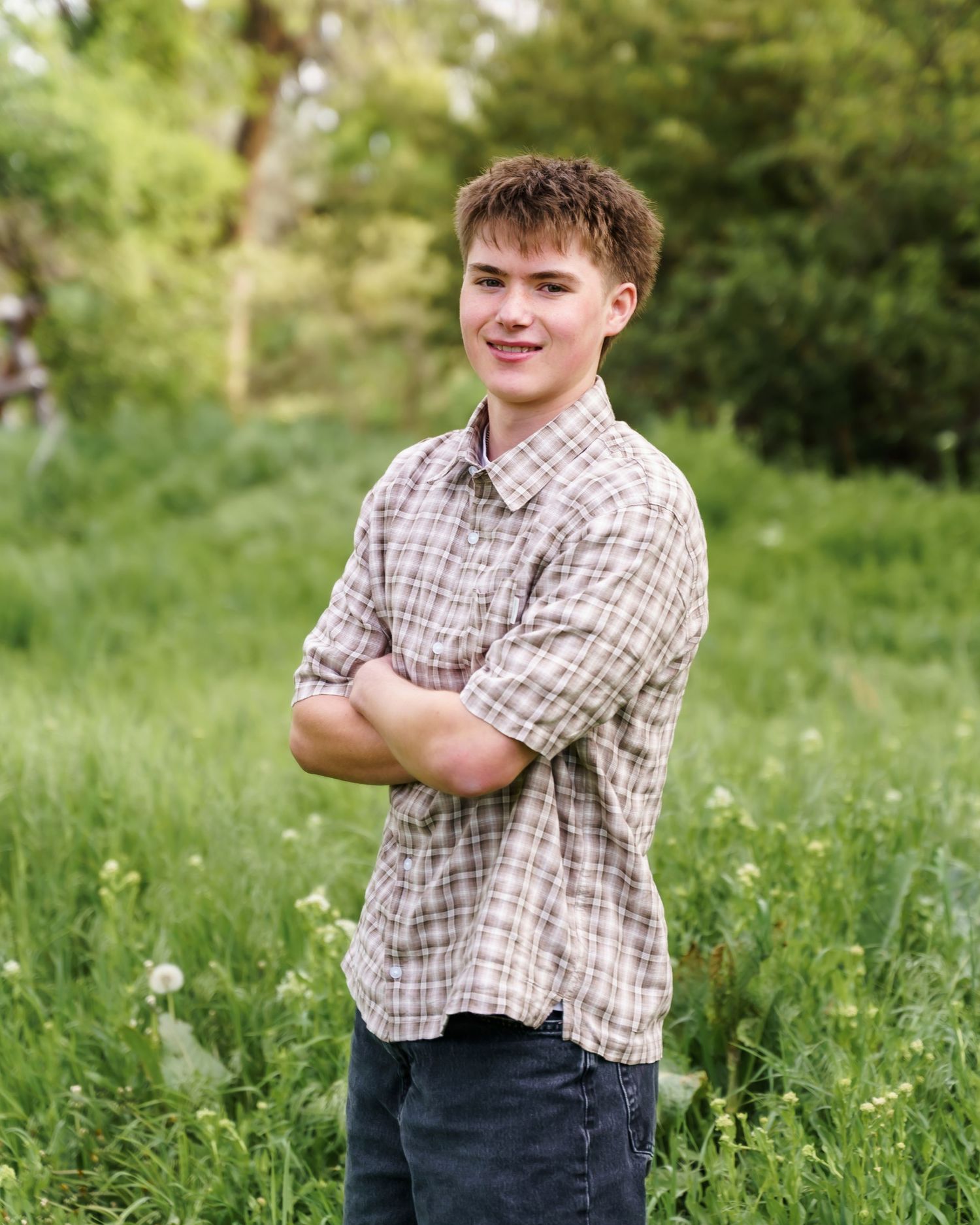 A smiling teenage boy in a plaid shirt stands with arms crossed in a lush green meadow for senior portraits.