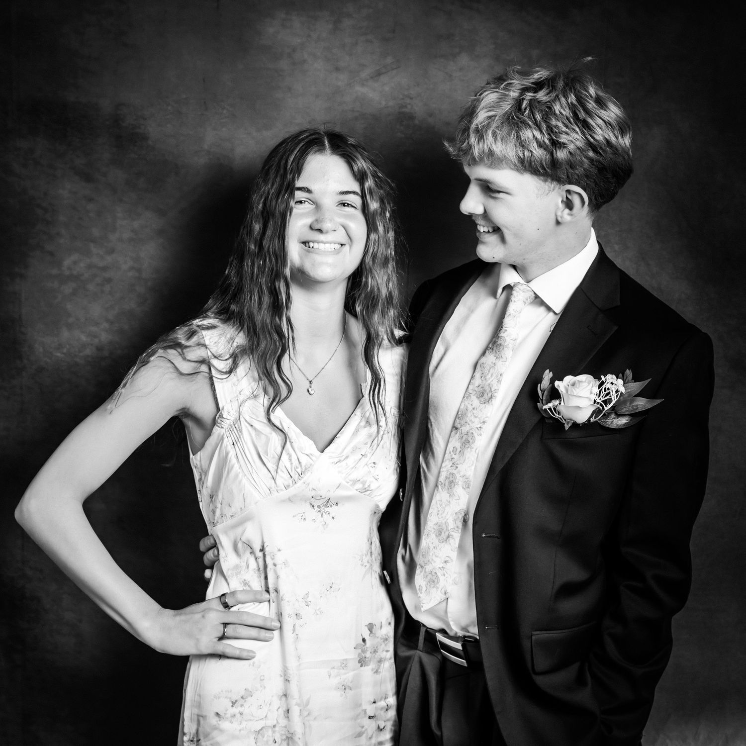 Black and white photo of a smiling young couple dressed formally, the woman in a floral dress and the man in a suit with boutonniere.