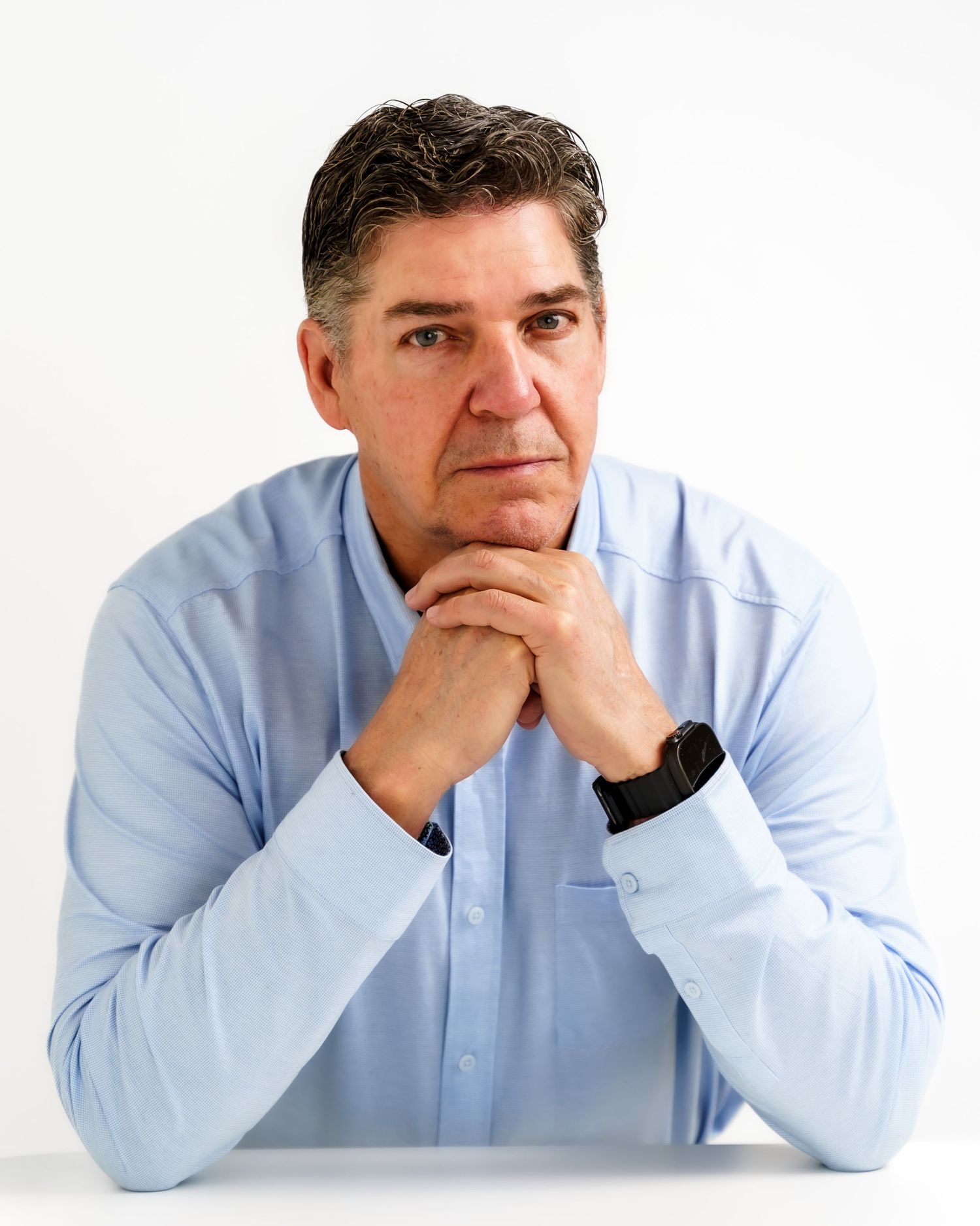 Middle-aged man in light blue shirt sits thoughtfully with hands clasped, posing against white background.