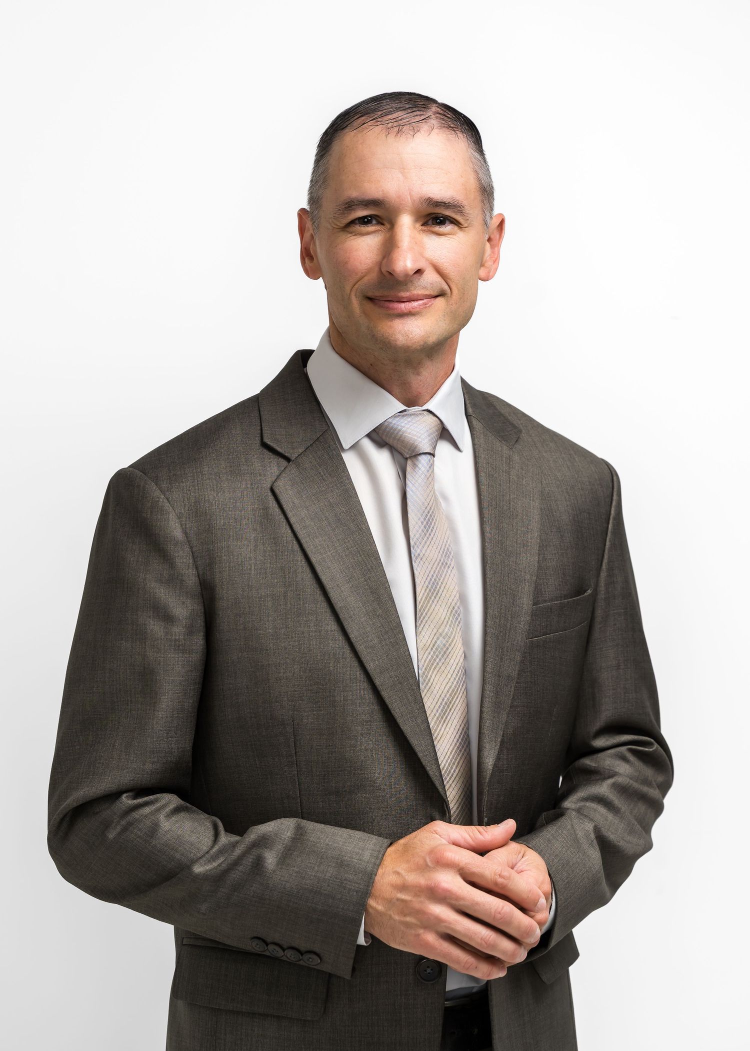 Professional businessman in gray suit and tie posing with hands clasped against white background.