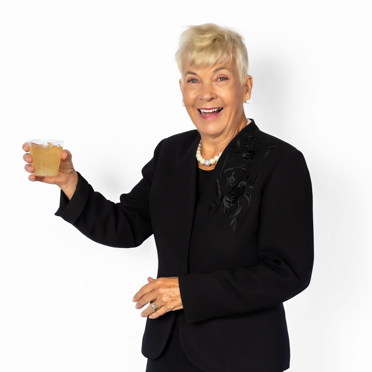 Smiling senior woman in black blazer holding a small wooden box, wearing pearl necklace, on white background.