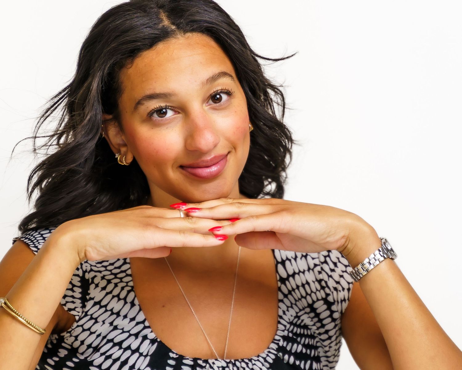 Smiling woman with dark wavy hair posing with hands clasped under chin, wearing patterned dress and gold jewelry.