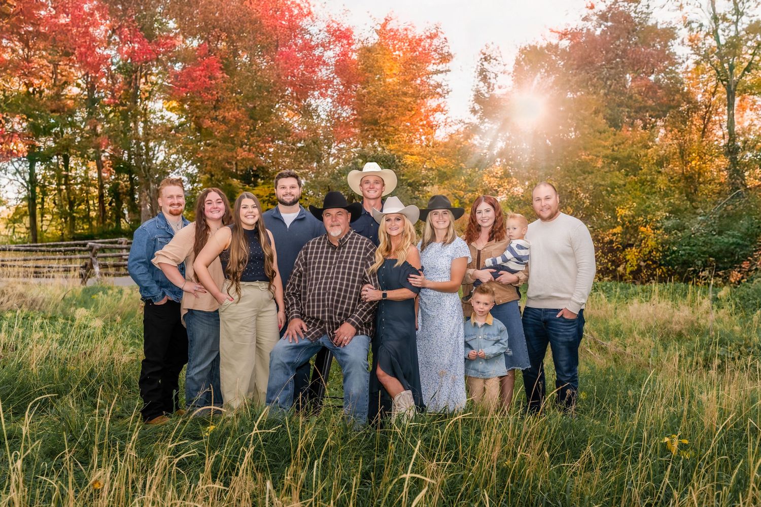 Family portrait of ten people posing outdoors in a field with vibrant fall foliage and golden sunlight.