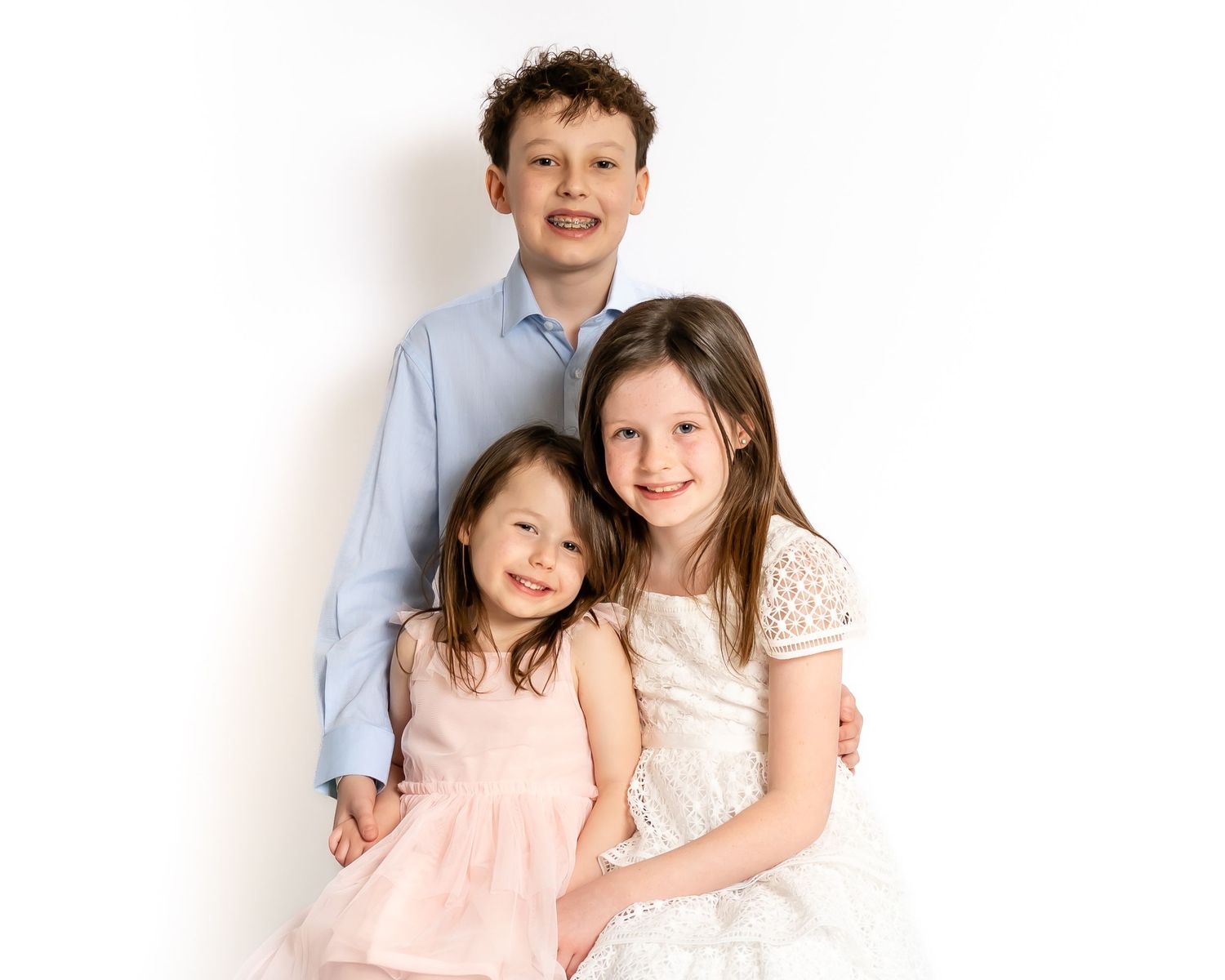 Three siblings posing together, older boy hugging two younger sisters in white studio portrait.