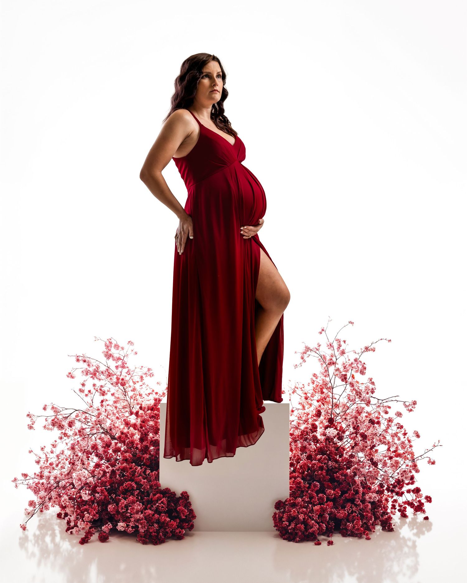 Elegant pregnant woman in flowing red gown standing on white pedestal surrounded by pink flowers.