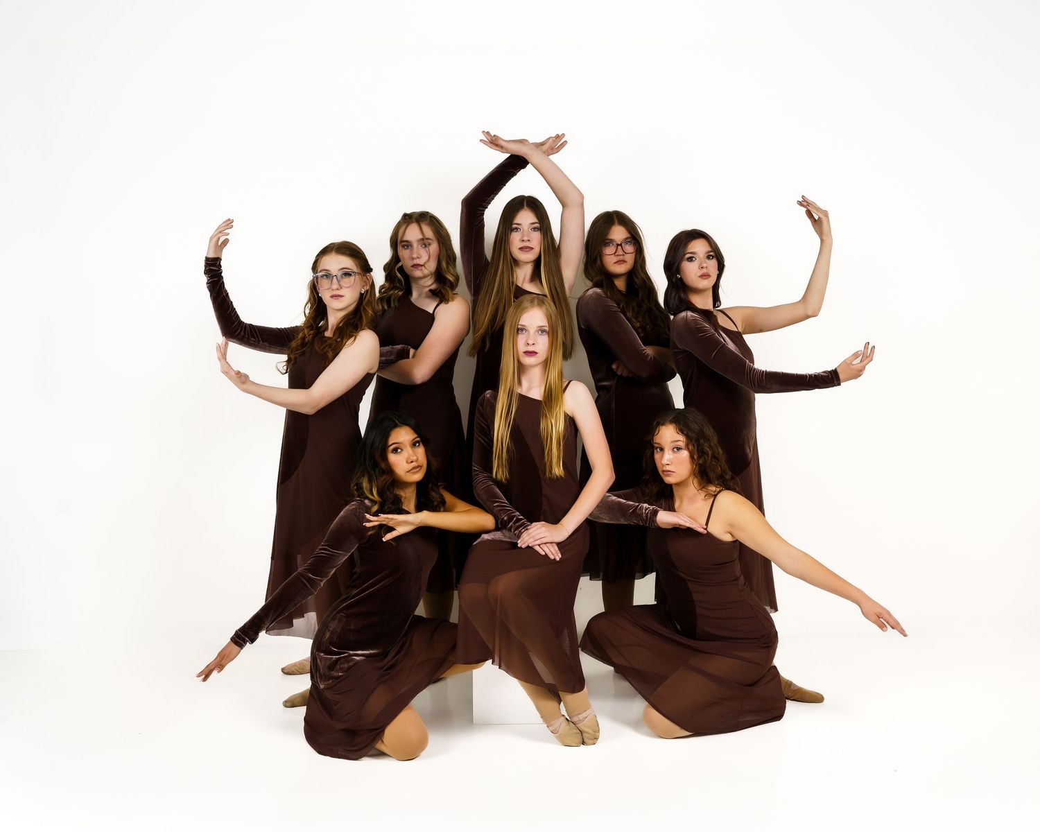 Group of nine women in coordinated brown dresses posing dynamically against white background in artistic dance formation.
