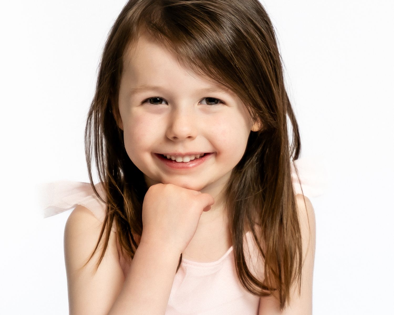 Adorable young girl in pink top smiles brightly with chin resting on fist in studio portrait.